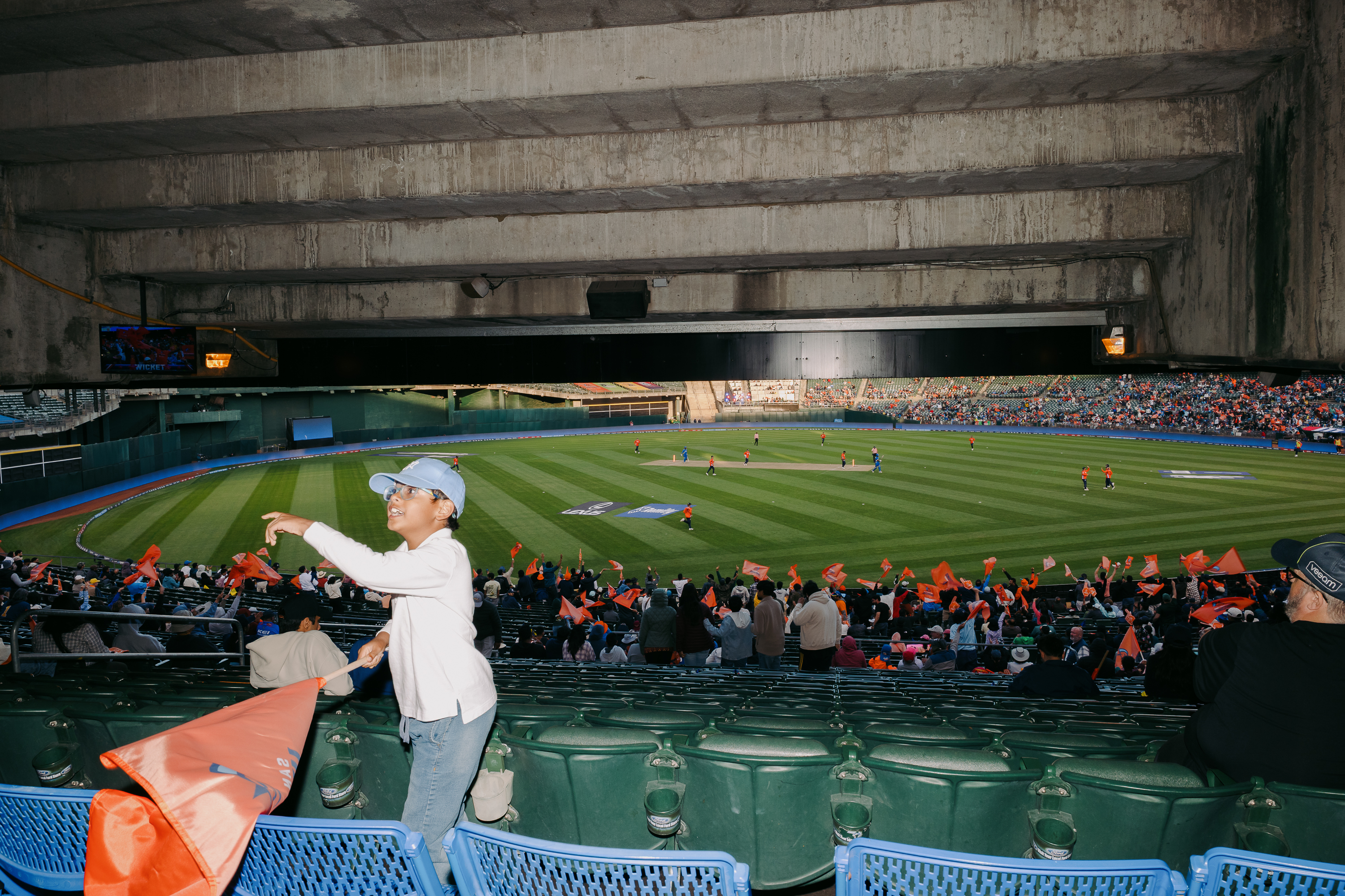 A child in a blue cap waves an orange flag in a stadium with green seats. In the background, a cricket field is active, and the crowd holds similar flags.
