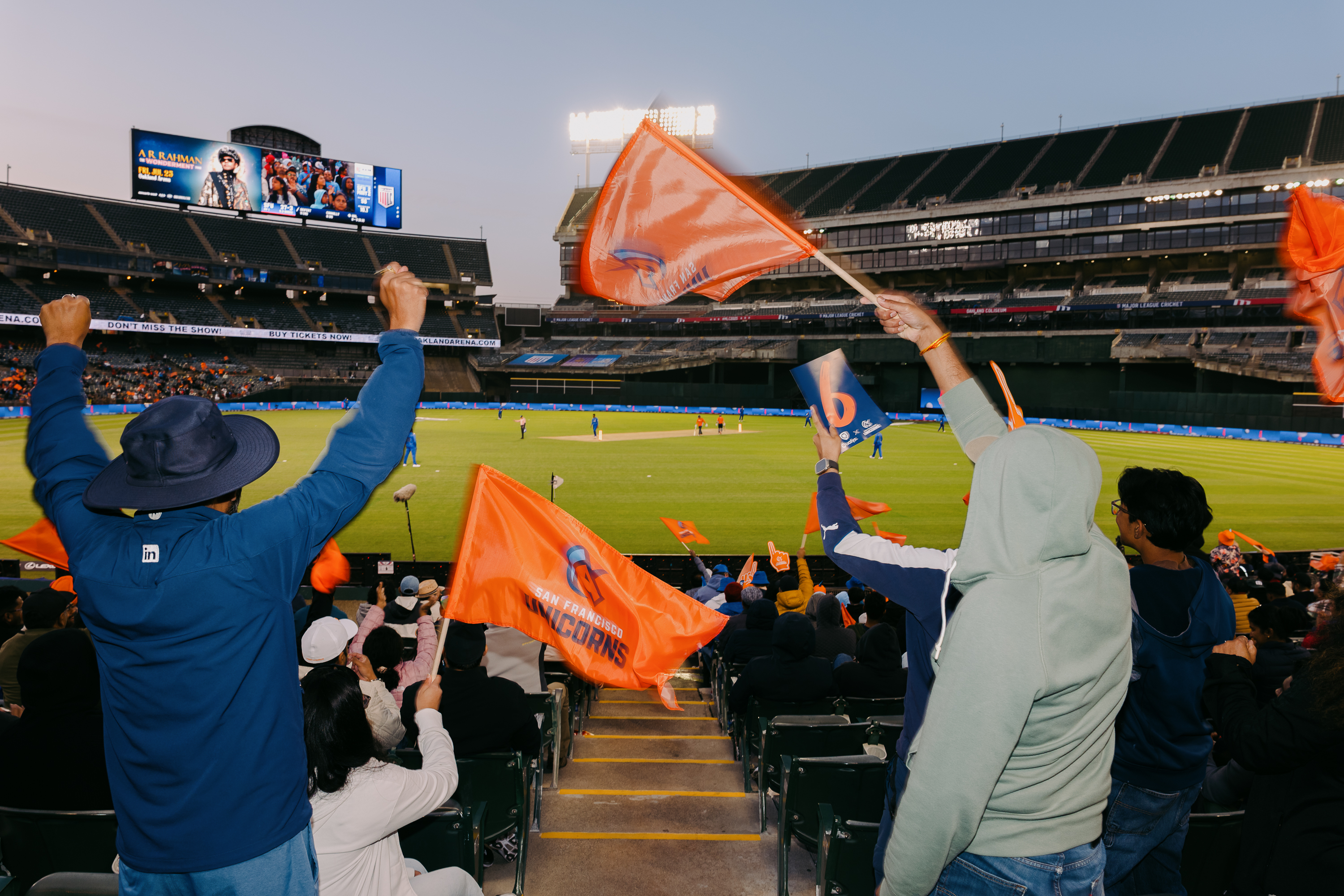 A crowd cheers at a cricket match in a large stadium. People wave orange flags with "San Francisco Unicorns" printed on them, creating a lively atmosphere.