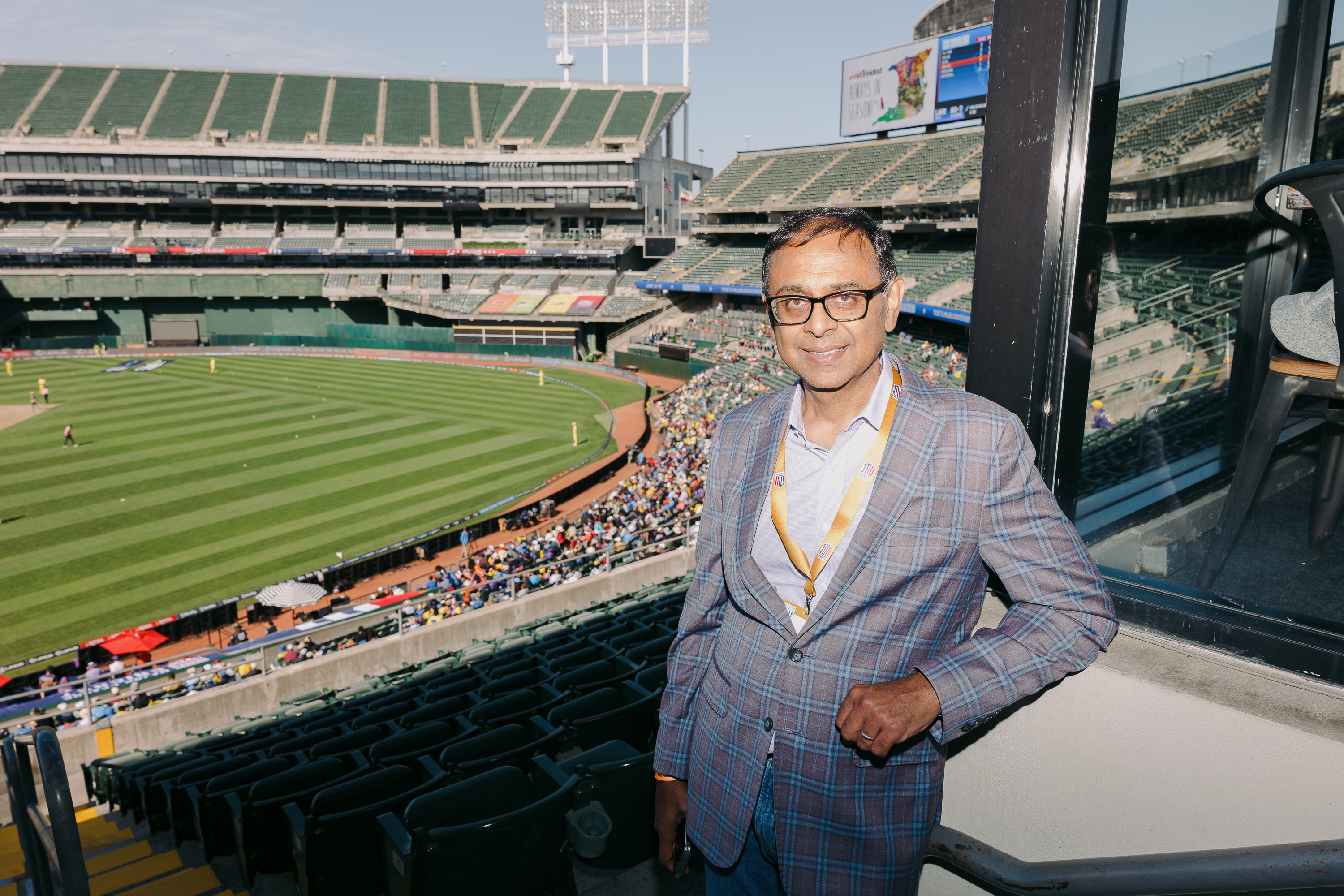 A man in a plaid jacket and glasses stands by a window overlooking a cricket stadium with green seats and a match in progress on the field.