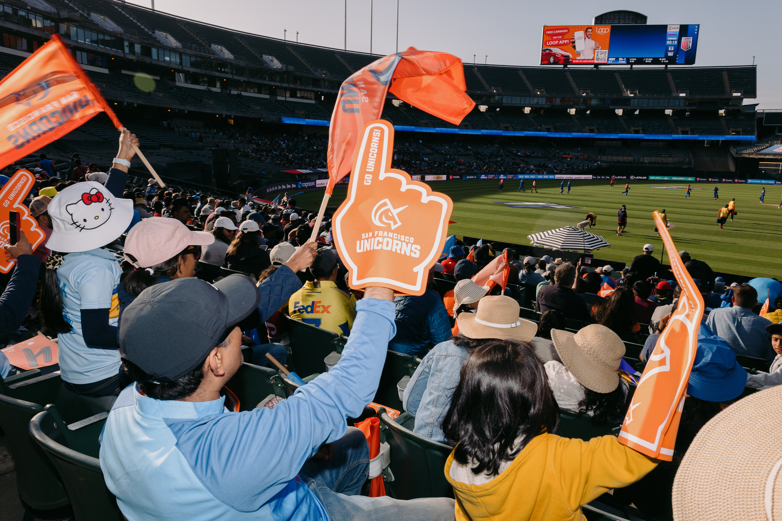 A lively stadium scene features cheering cricket fans with orange flags and foam fingers reading &quot;Go Unicorns!&quot; The match is visible on a sunny day.