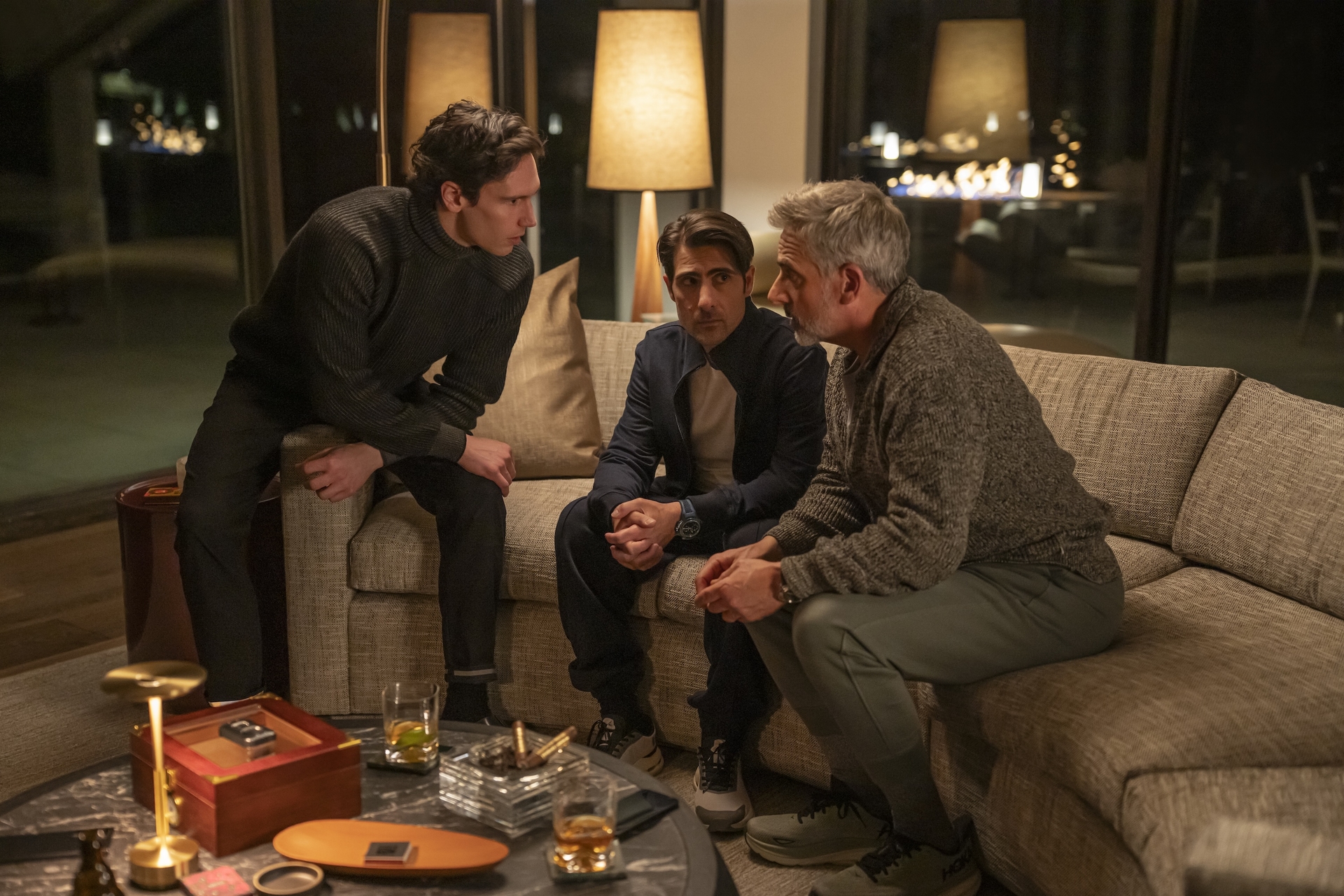 Three men are having a serious conversation in a dimly lit living room, seated around a table with drinks and a lamp.