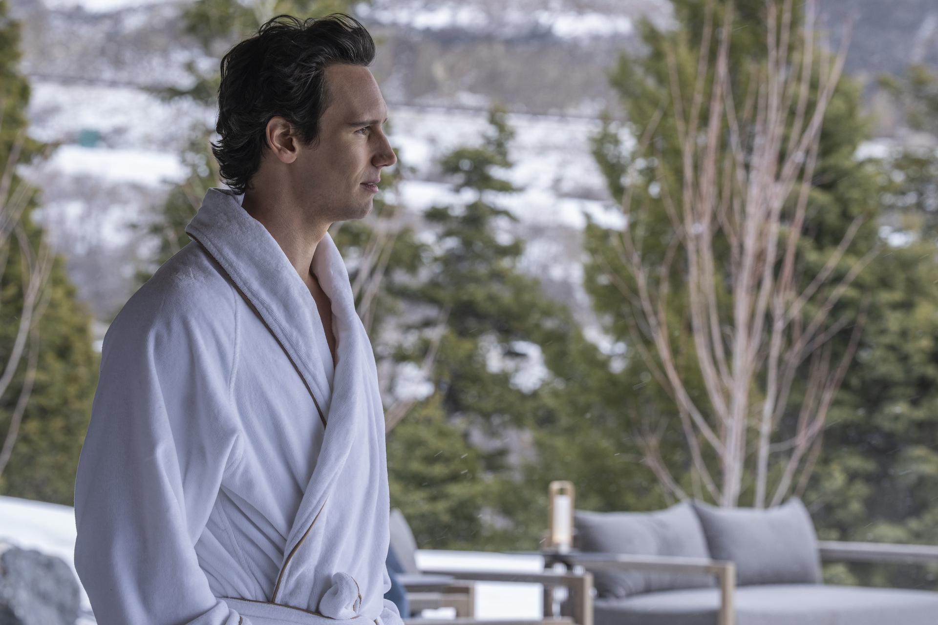 A person in a white bathrobe stands outdoors, surrounded by snow-covered trees. Behind them is a modern outdoor couch with a blurred wintry background.