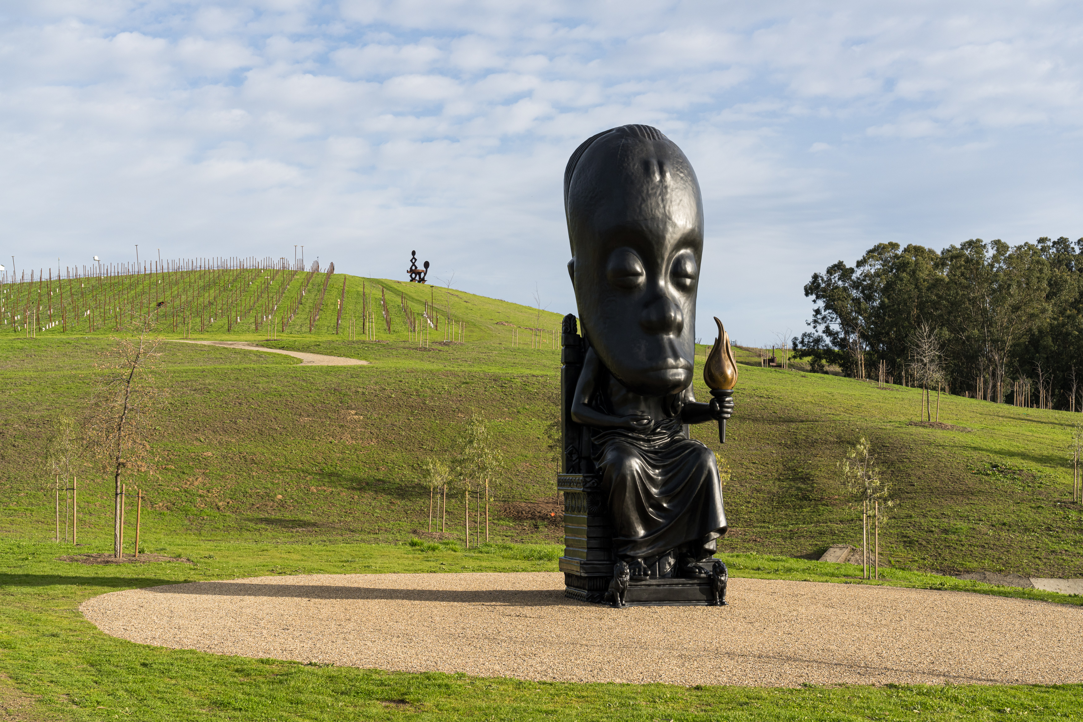 A large, black statue with an oversized head sits on gravel, holding a torch. It's set in a grassy, hilly landscape with trees and a vineyard in the background.