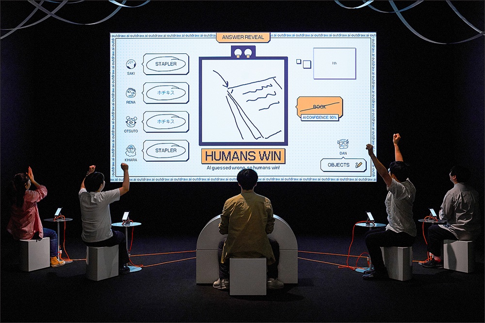 Five people sit in front of a large screen displaying a drawing game where humans won against AI. They're raising their fists in celebration.