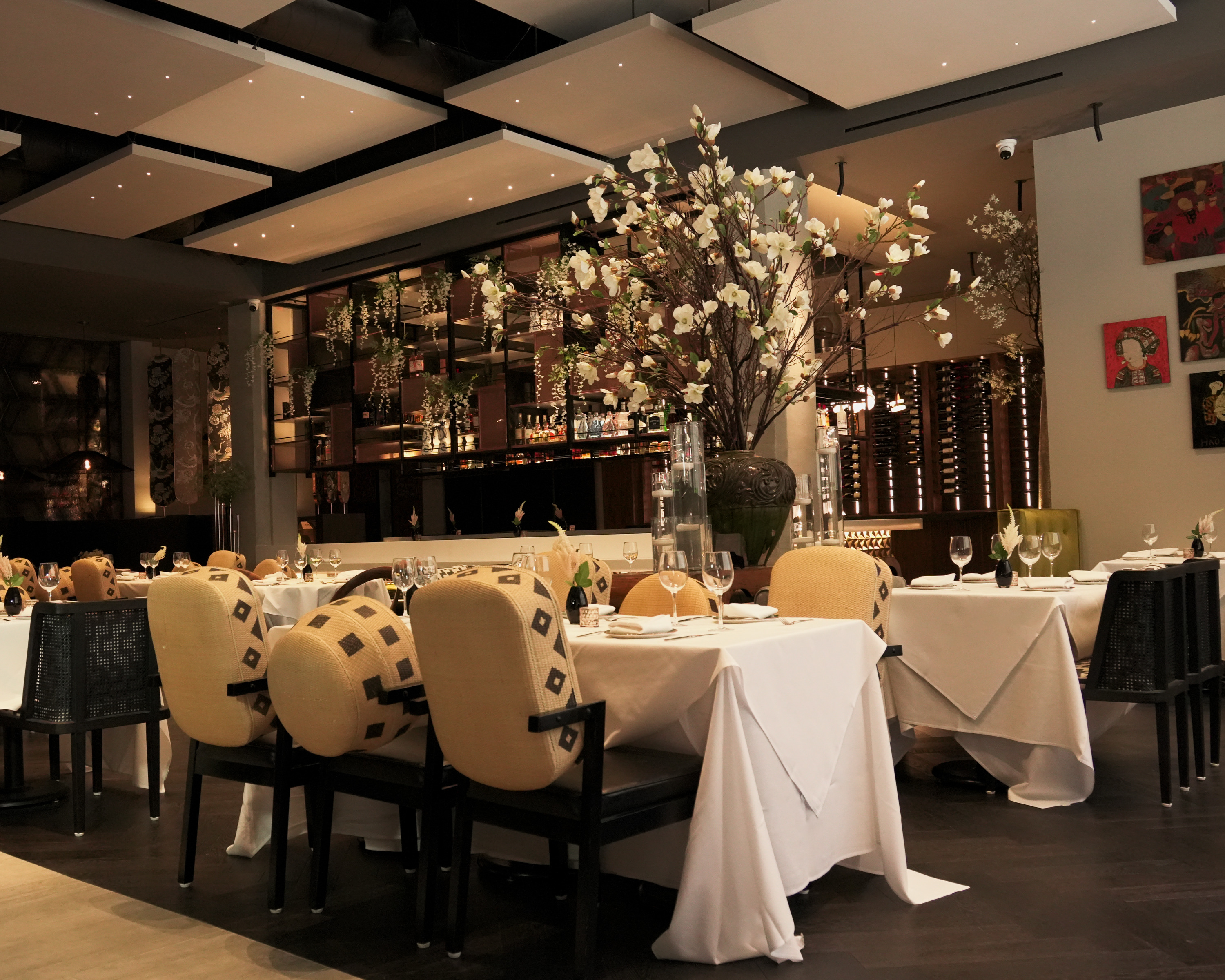 The image shows an elegant restaurant with neatly set tables, cushioned chairs, and a large floral centerpiece. There are decorative art pieces on the walls.