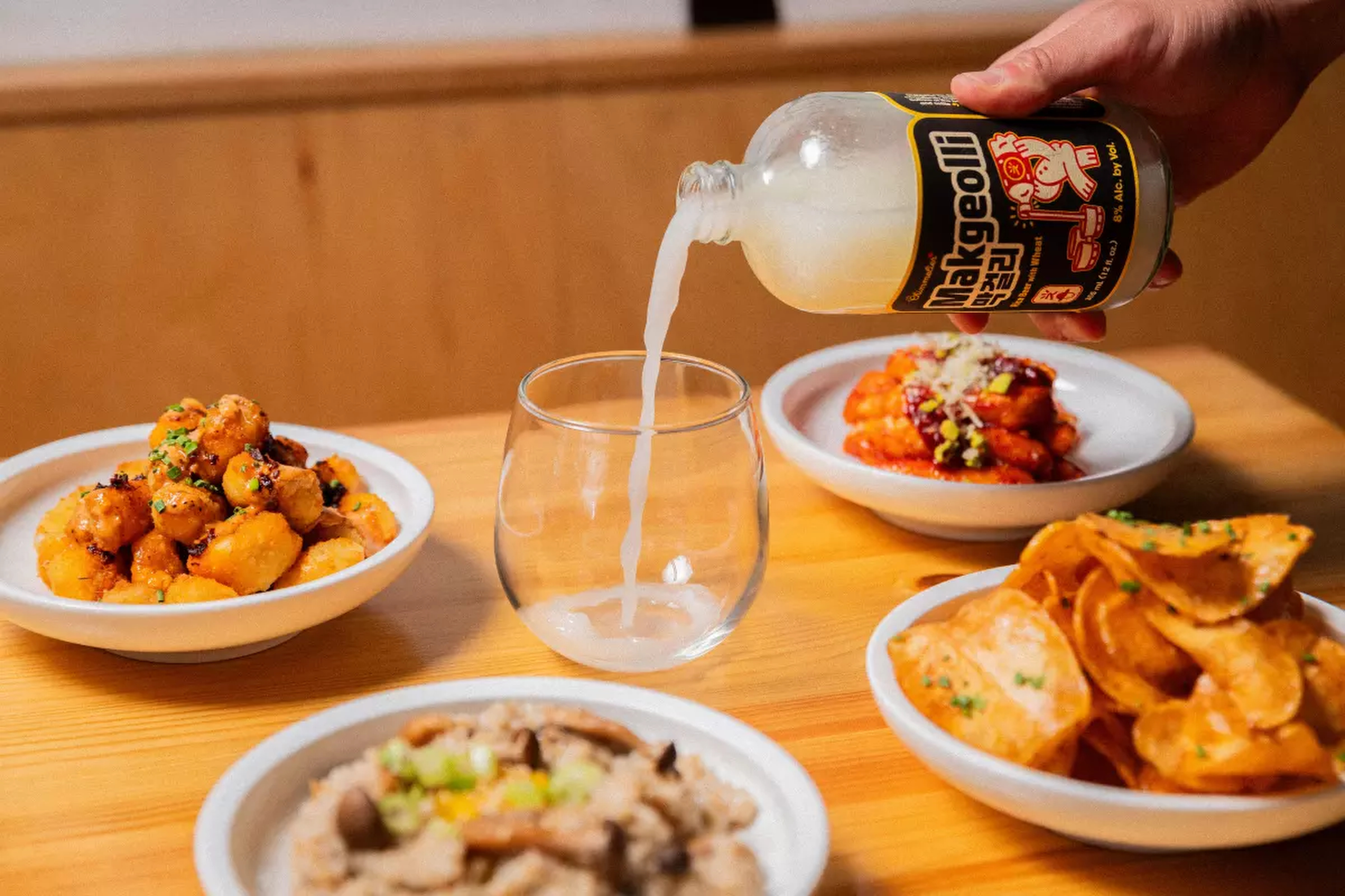A hand pours a cloudy beverage from a bottle into a glass. Surrounding the glass are bowls of various dishes, including crispy snacks and sauced items.