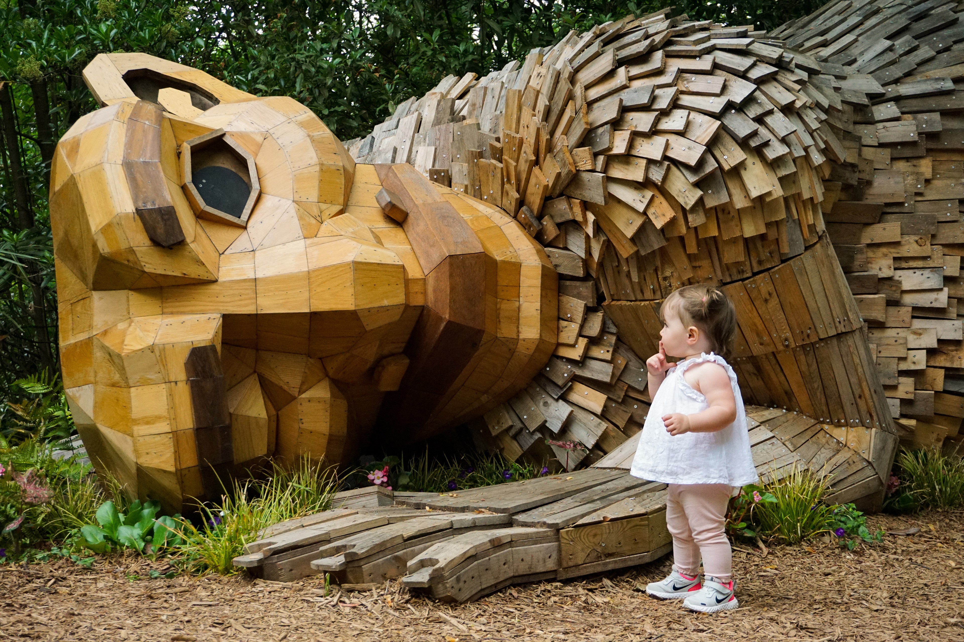 A small child stands in front of a large wooden sculpture of a bear's head, surrounded by greenery. The child appears curious, with a hand near their mouth.