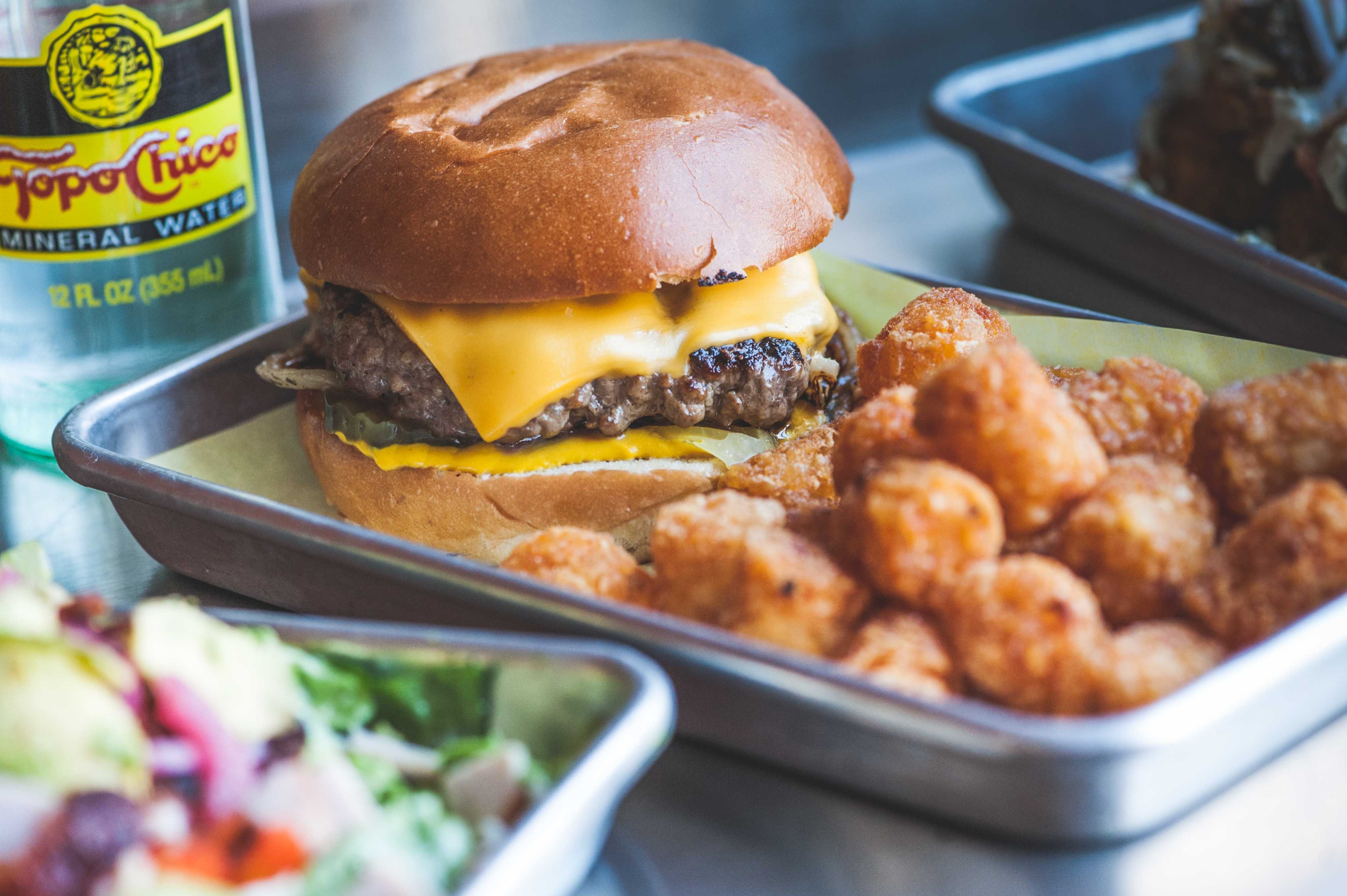 A cheeseburger with a beef patty and melted cheese sits on a tray next to crispy tater tots, accompanied by a bottle of Topo Chico mineral water.