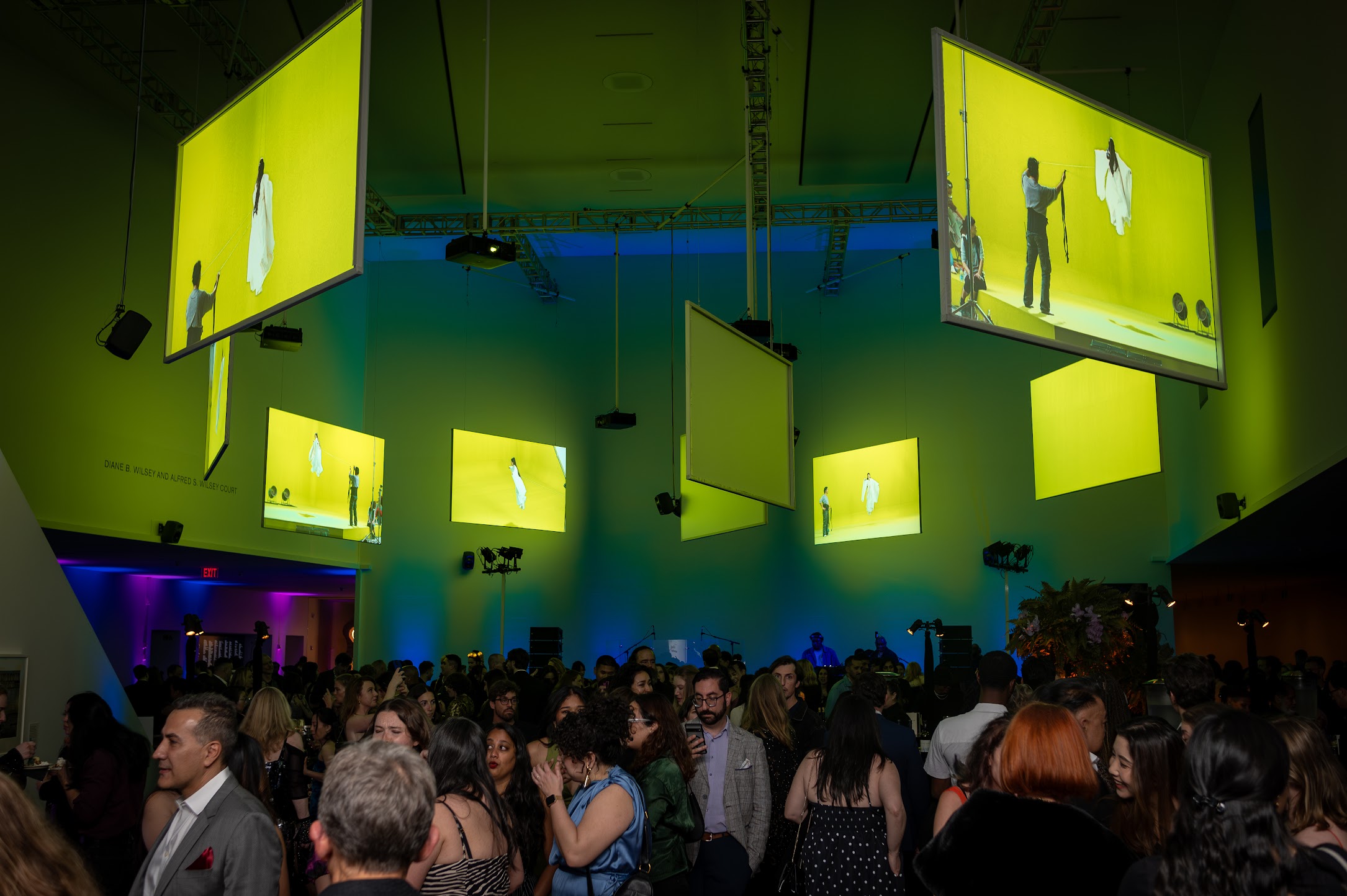 A crowded room with people mingling under hanging screens displaying images on a bright yellow backdrop. The ambiance is lively with vibrant lighting.