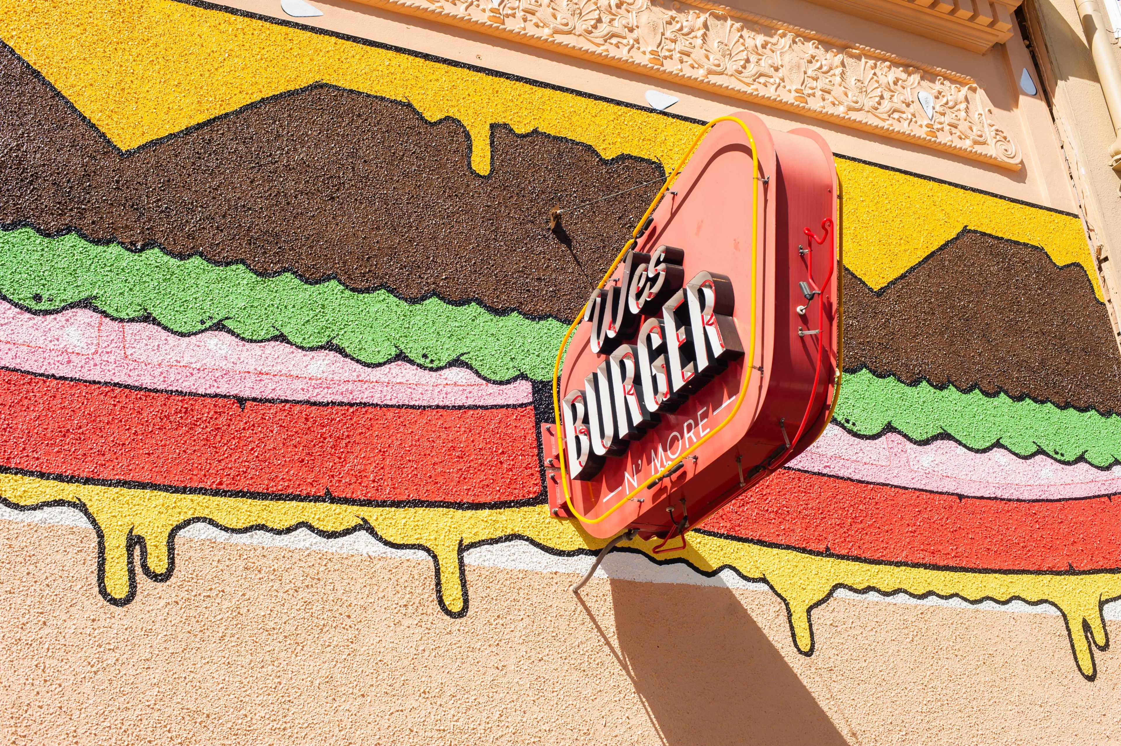 A colorful mural depicts a giant burger with layers of toppings on a wall. A red and yellow neon sign reads &quot;Wes Burger 'N' More,&quot; protruding from the mural.