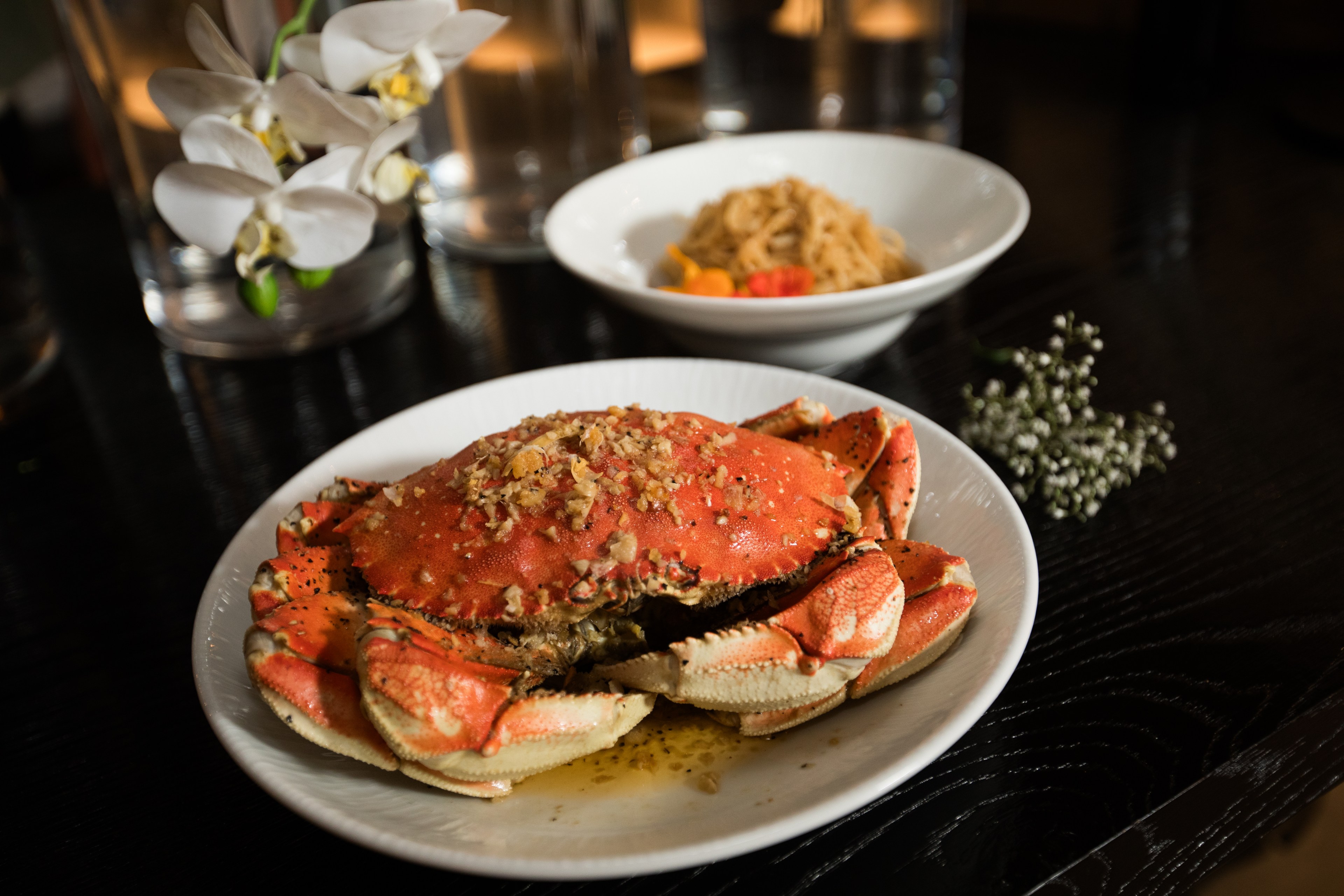 A cooked crab with herbs is on a white plate, accompanied by a bowl of noodles. White orchids and a small flower bunch are nearby on a dark table.