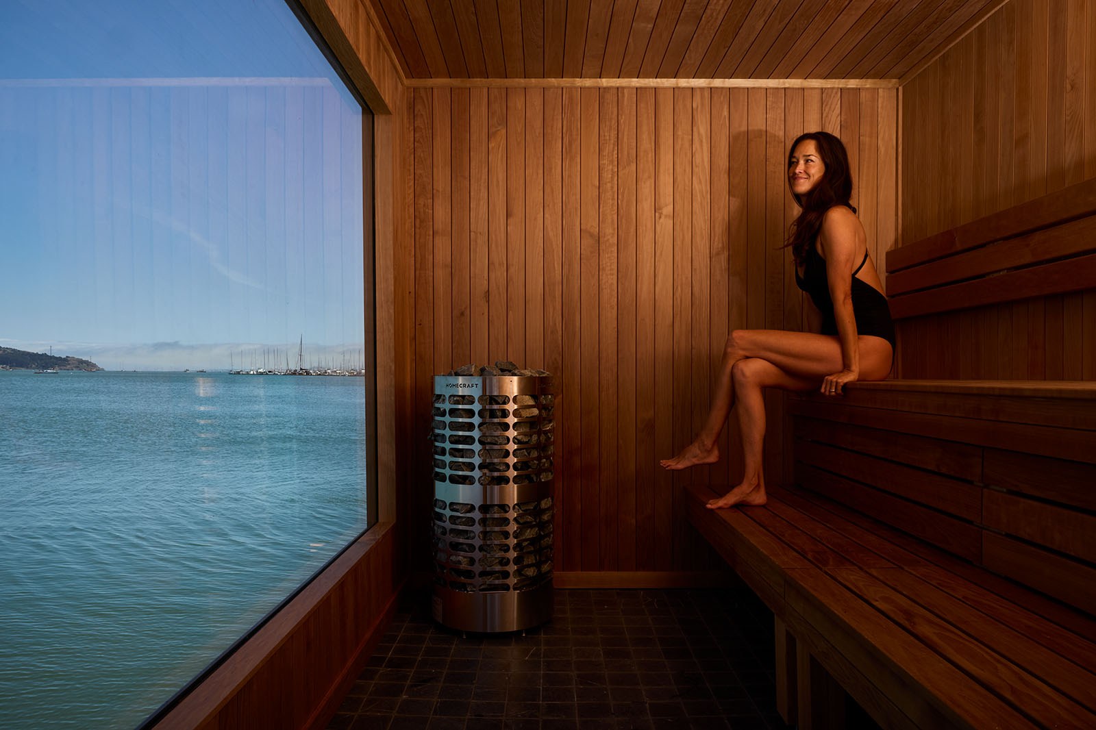 A woman in a black swimsuit sits on a wooden bench in a sauna with a large window offering a scenic view of a calm body of water and distant hills.
