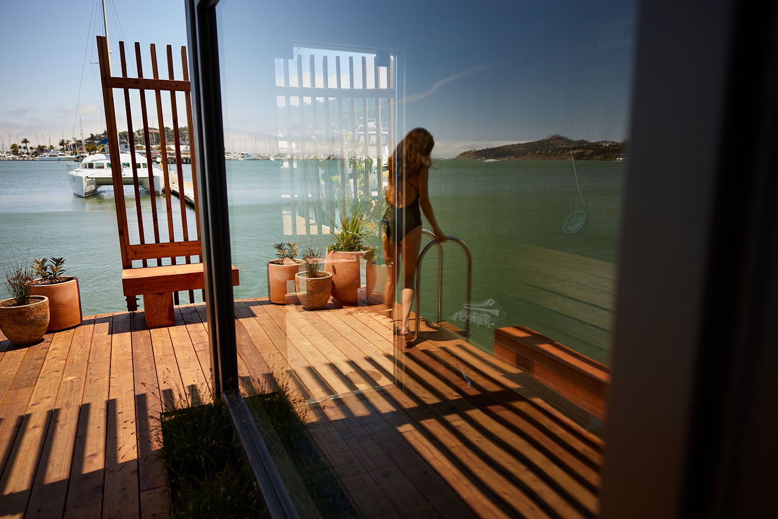 A serene dock scene shows a person on a wooden deck by calm water, with potted plants and yachts in the background, all reflected in a glass surface.