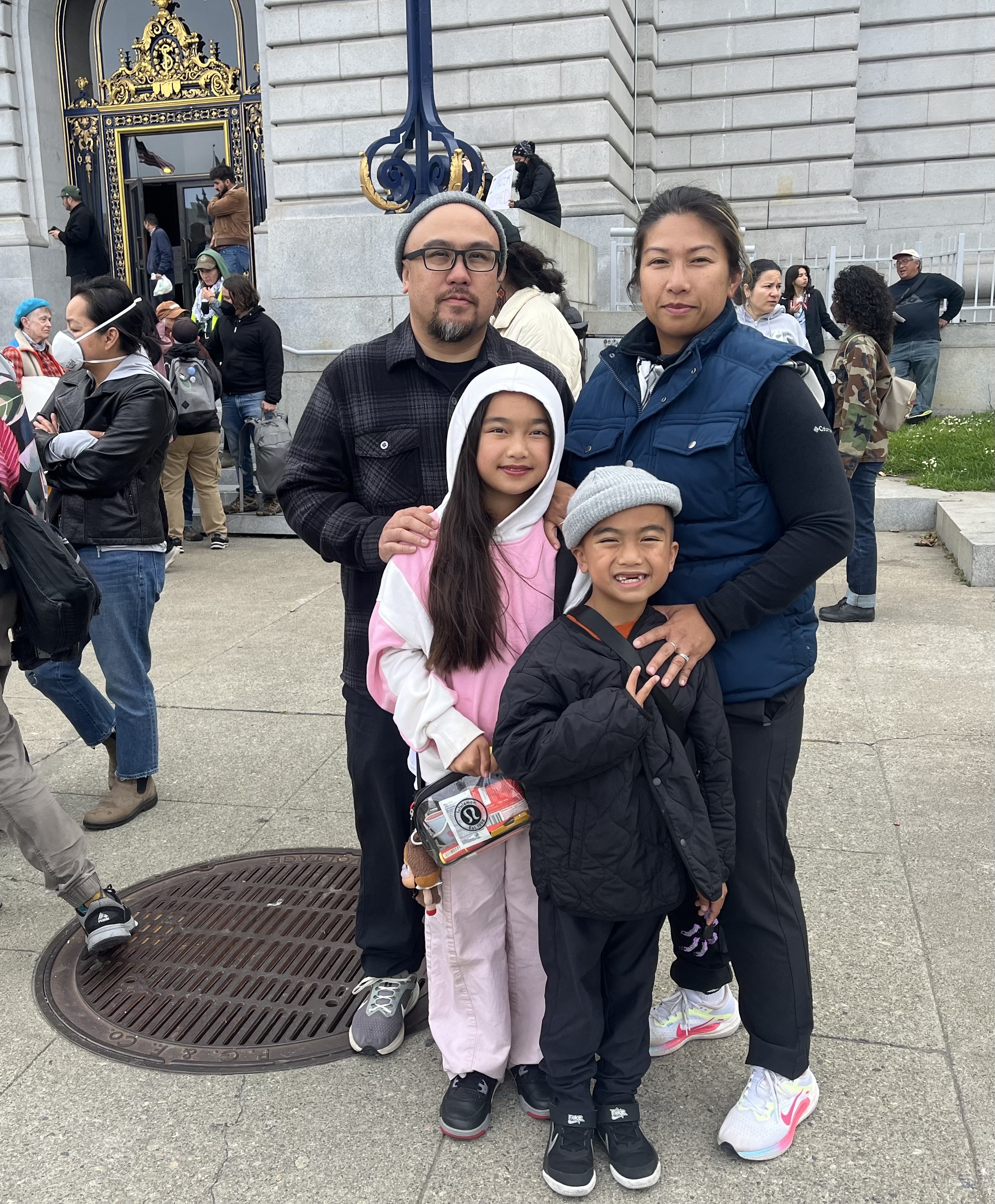 A family of four is posing outside a grand building with ornate details. They are dressed warmly, and there are people and activity in the background.