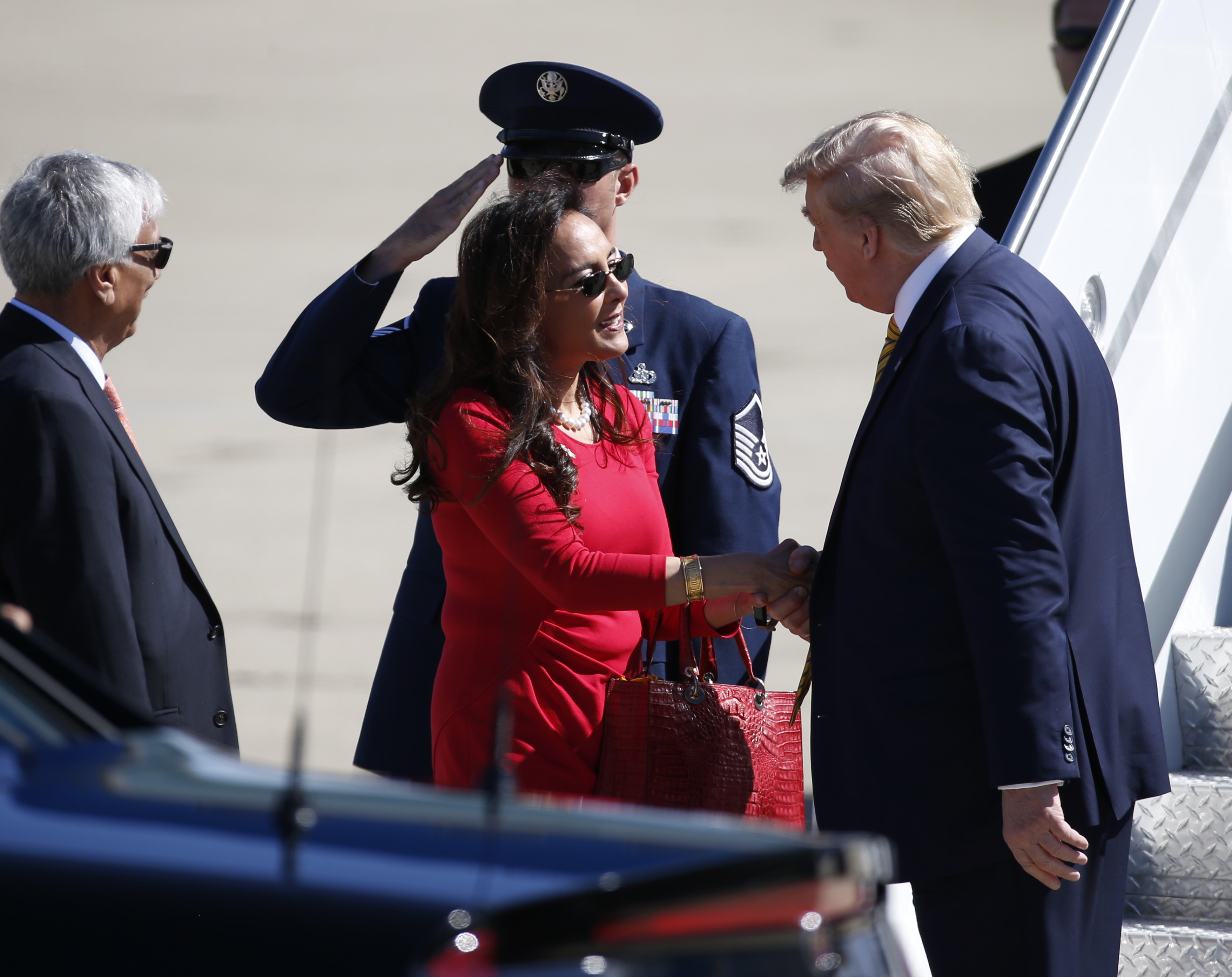 A woman in red shakes hands with a man in a suit near an airplane staircase. A man in a military uniform salutes, and another person stands nearby.