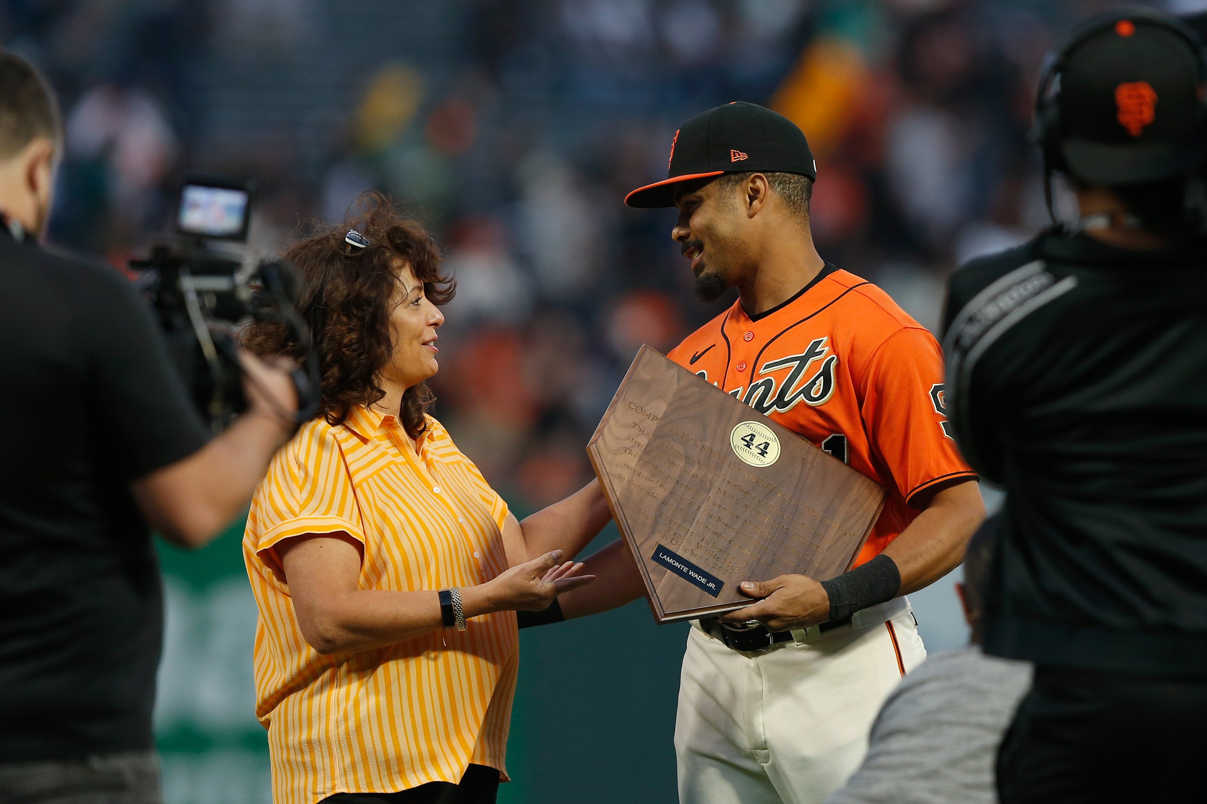 A woman in a yellow shirt hands a wooden plaque to a baseball player in an orange Giants jersey. A cameraman records the moment.