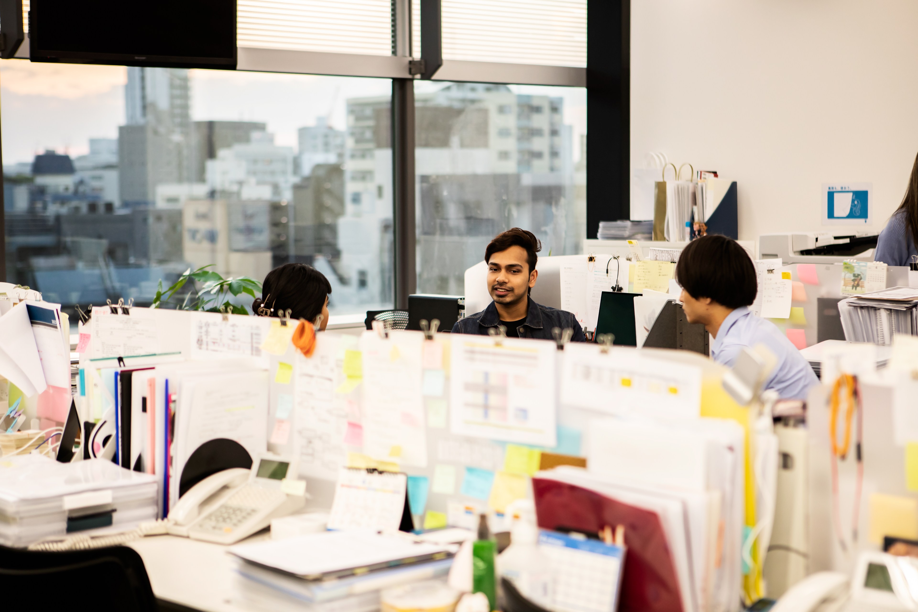 Three people are talking in a bright, cluttered office. The room is filled with papers, folders, and computers. Large windows show a cityscape outside.