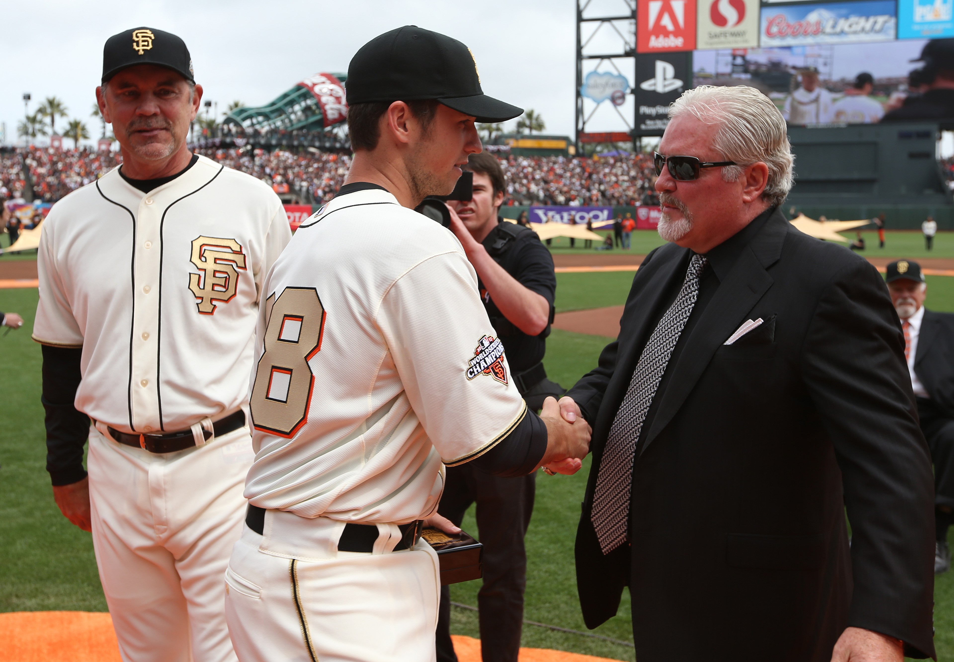 A baseball player in a white Giants uniform shakes hands with a man in a suit and sunglasses. Another person in a Giants uniform stands nearby.