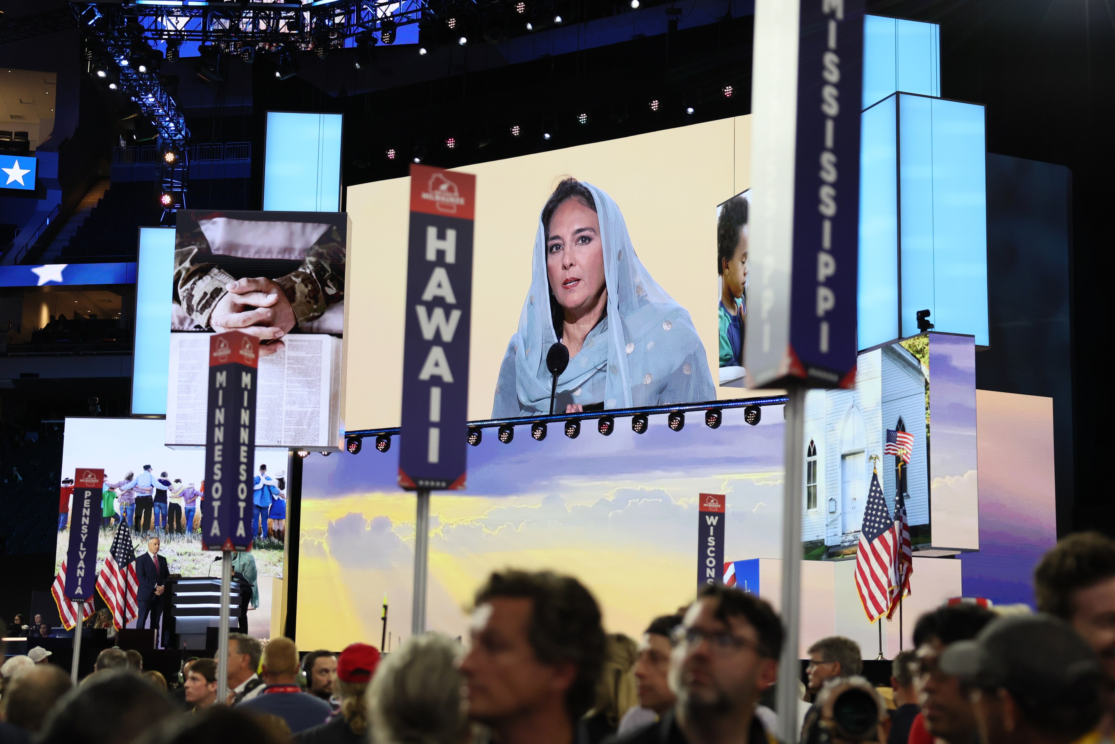 A woman in a blue headscarf speaks on a large screen at a crowded event. State signs like &quot;Hawaii&quot; and &quot;Mississippi&quot; rise above the audience.