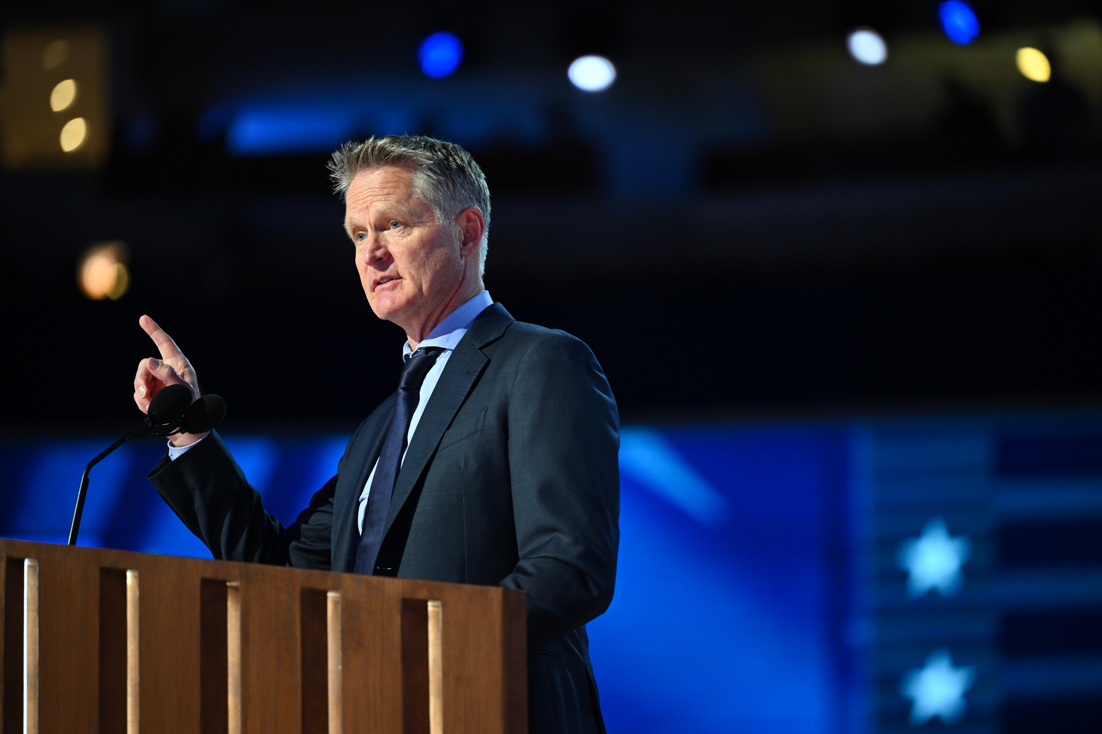 A man in a suit gestures while speaking at a wooden podium with a microphone, set against a blue-toned, blurred background featuring stars.