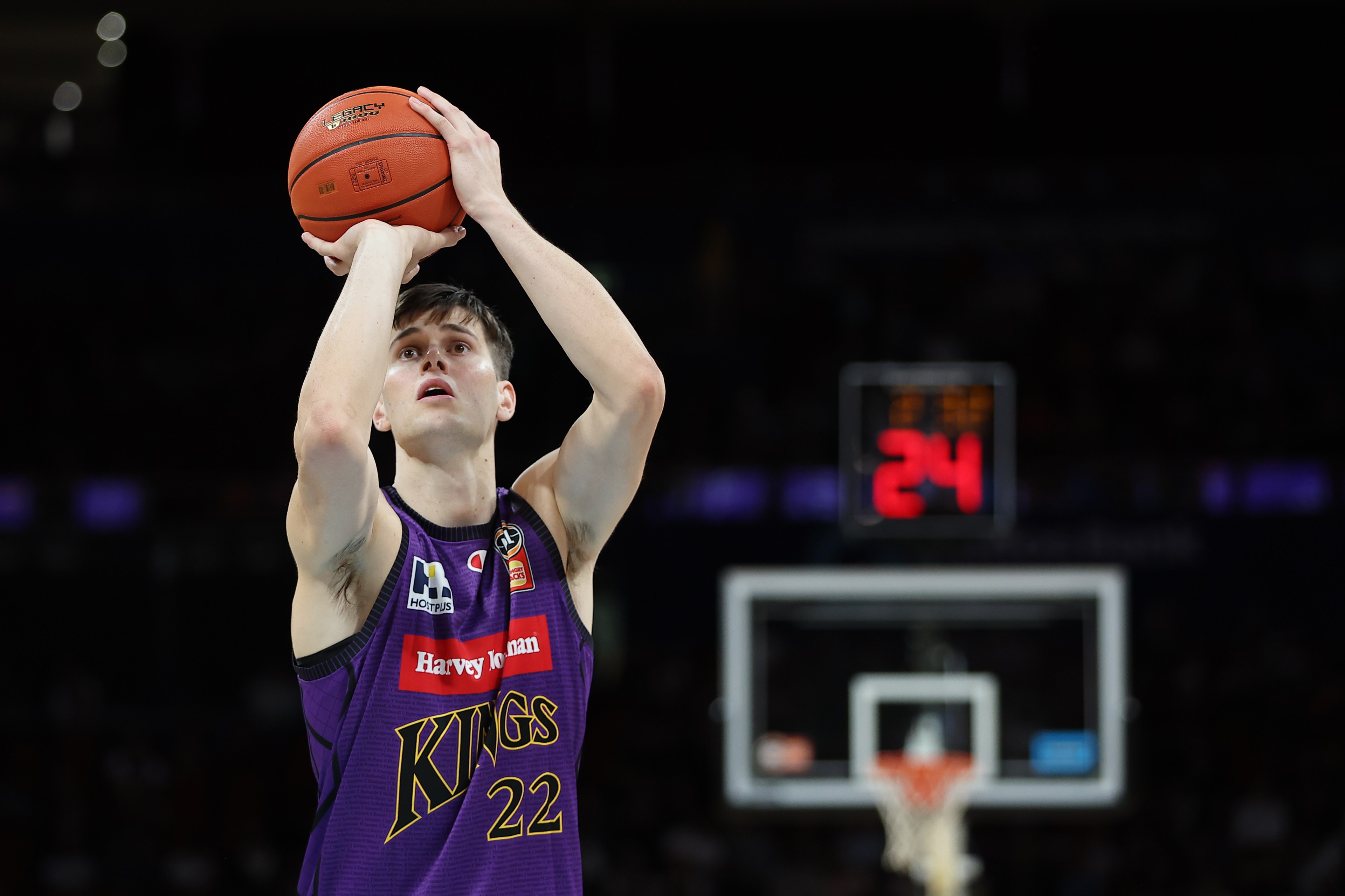 A basketball player in a purple "Kings" jersey aims a shot, with a ball above his head. The focus is sharp, while the background shows a blurred basketball hoop and scoreboard.