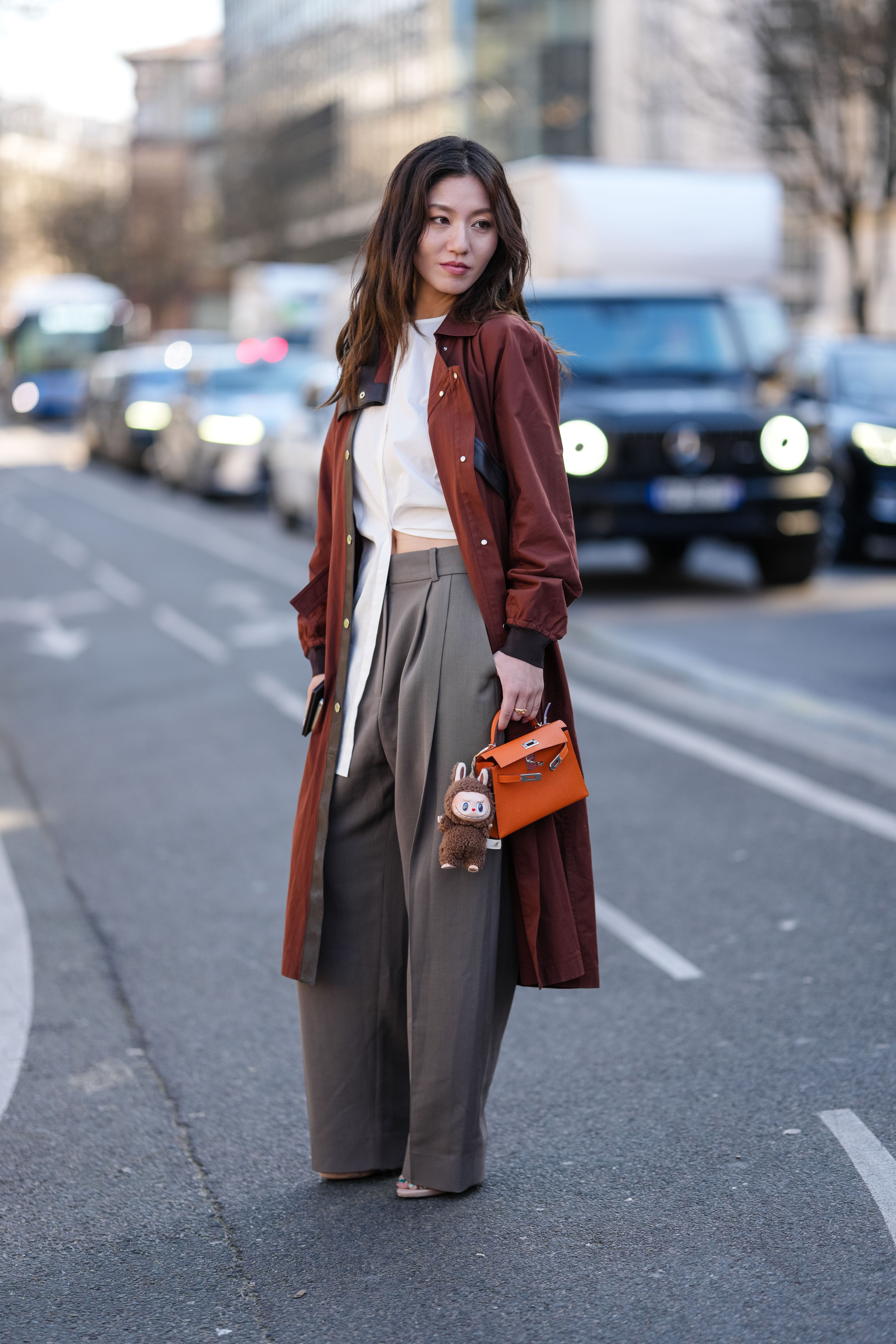 A woman stands on a city street wearing a long brown coat, white top, and gray pants. She holds a small orange handbag with a toy keychain. Cars are in the background.