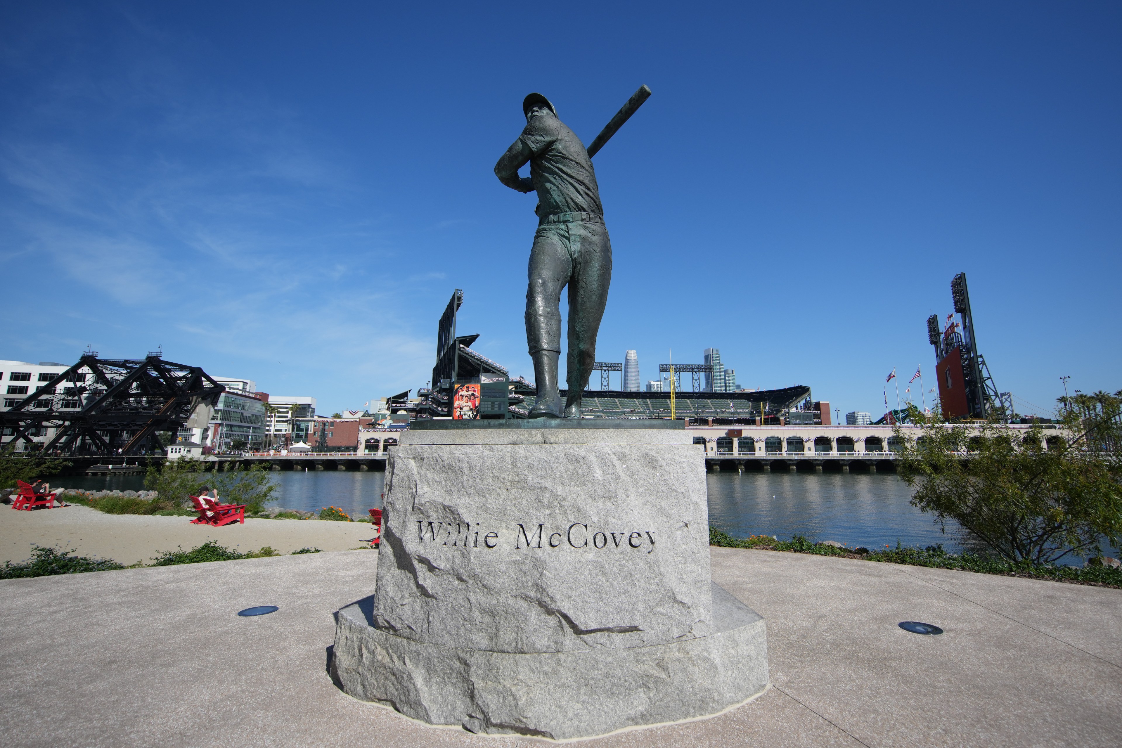 The image shows a statue of a baseball player swinging a bat, mounted on a stone pedestal with the name &quot;Willie McCovey.&quot; A stadium and river are in the background.