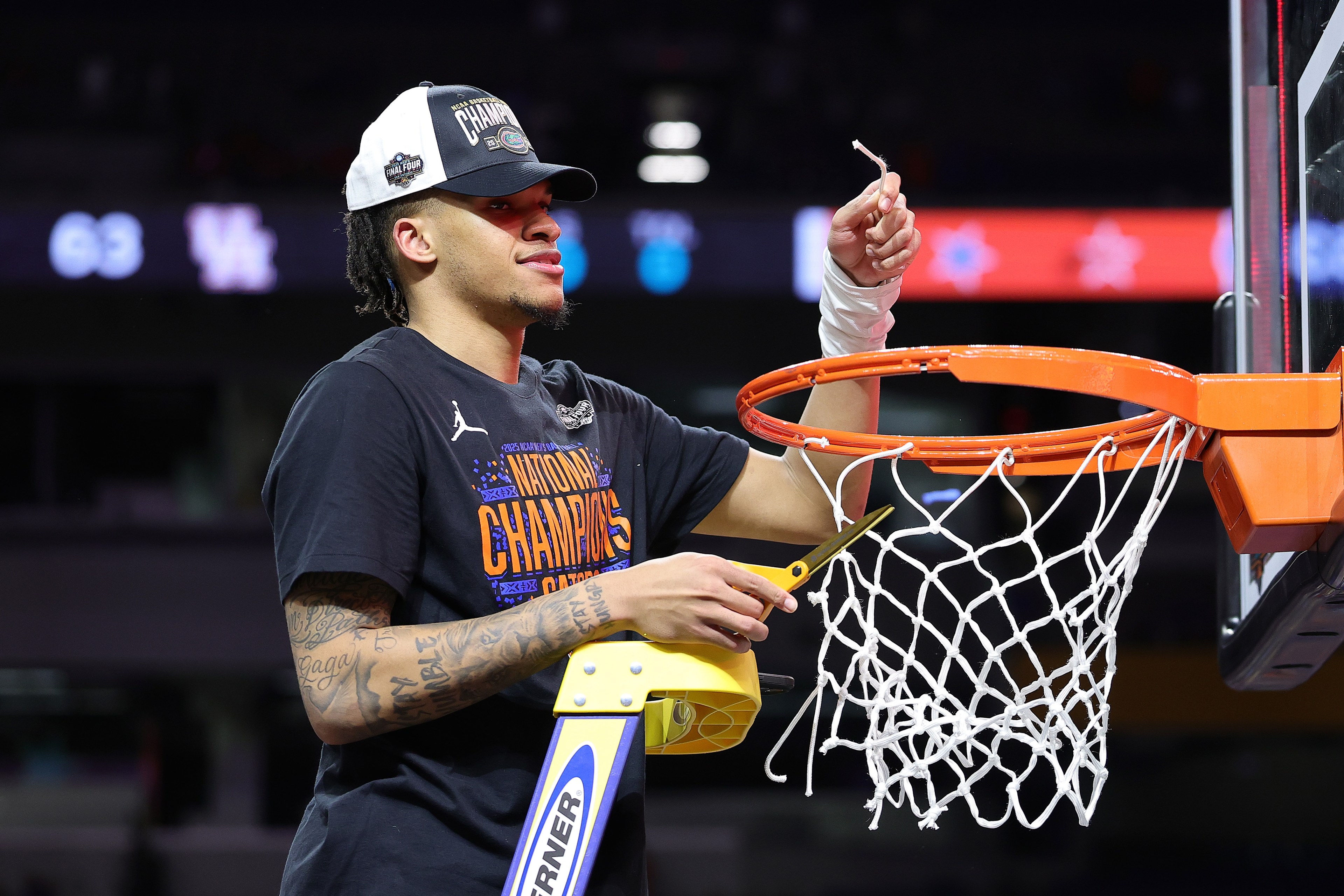 A person wearing a &quot;National Champions&quot; shirt and cap is cutting down a basketball net from the hoop, standing on a ladder while holding scissors.