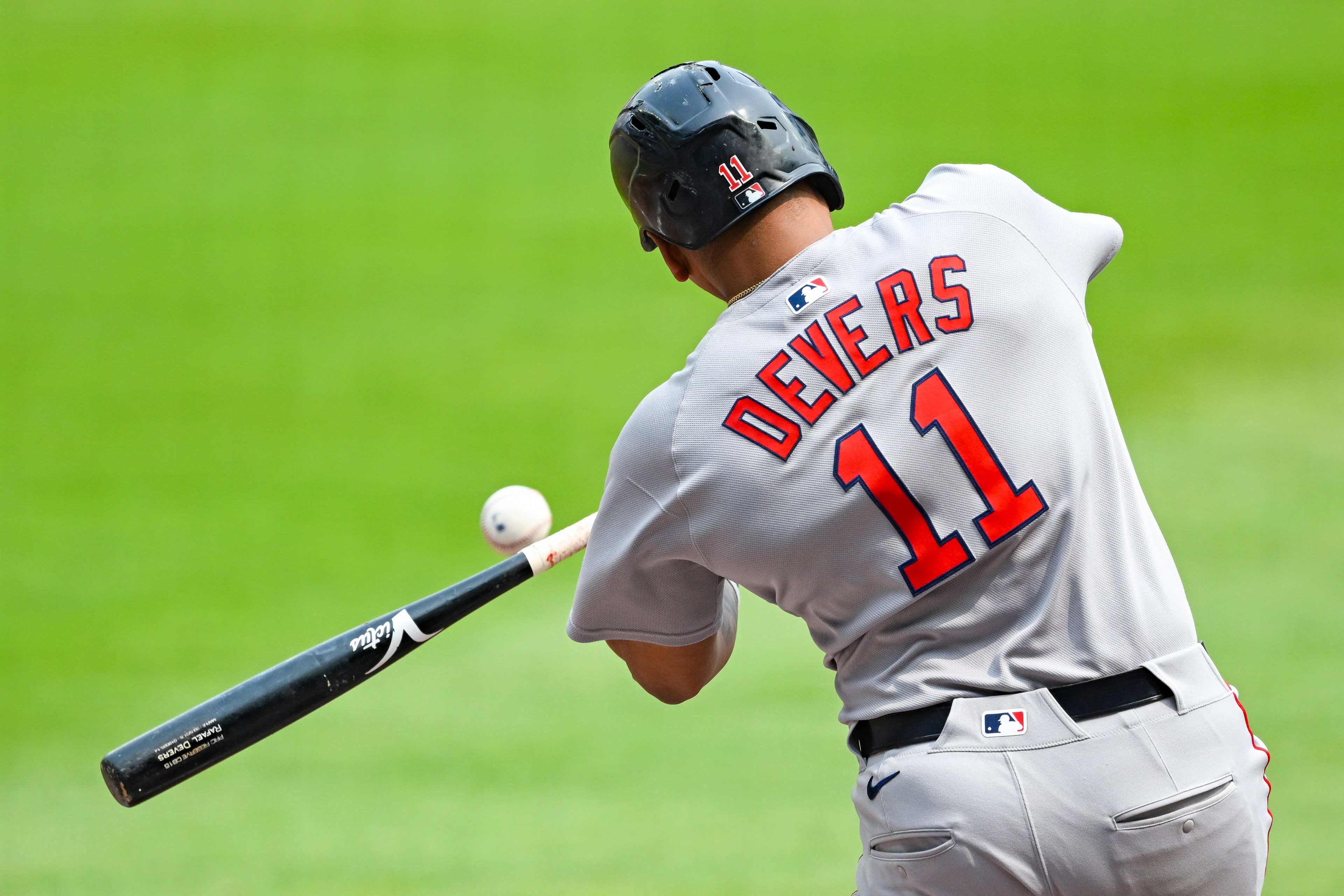 A baseball player in a gray uniform, with "Devers" and number 11 on the back, swings his bat at a fast-approaching ball. The background is a bright green field.