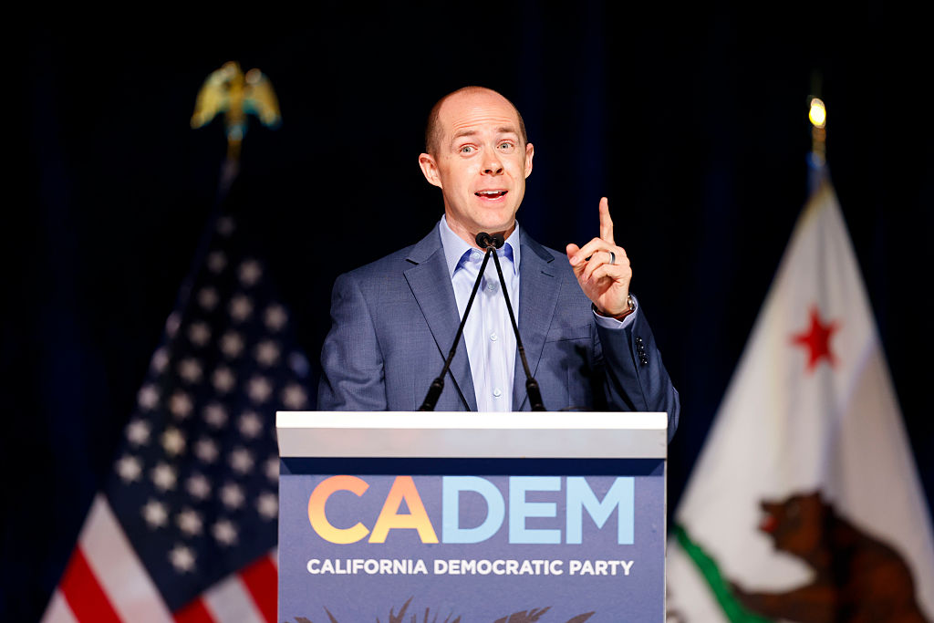 A man in a suit speaks passionately at a podium labeled &quot;CADEM California Democratic Party,&quot; with U.S. and California flags in the background.