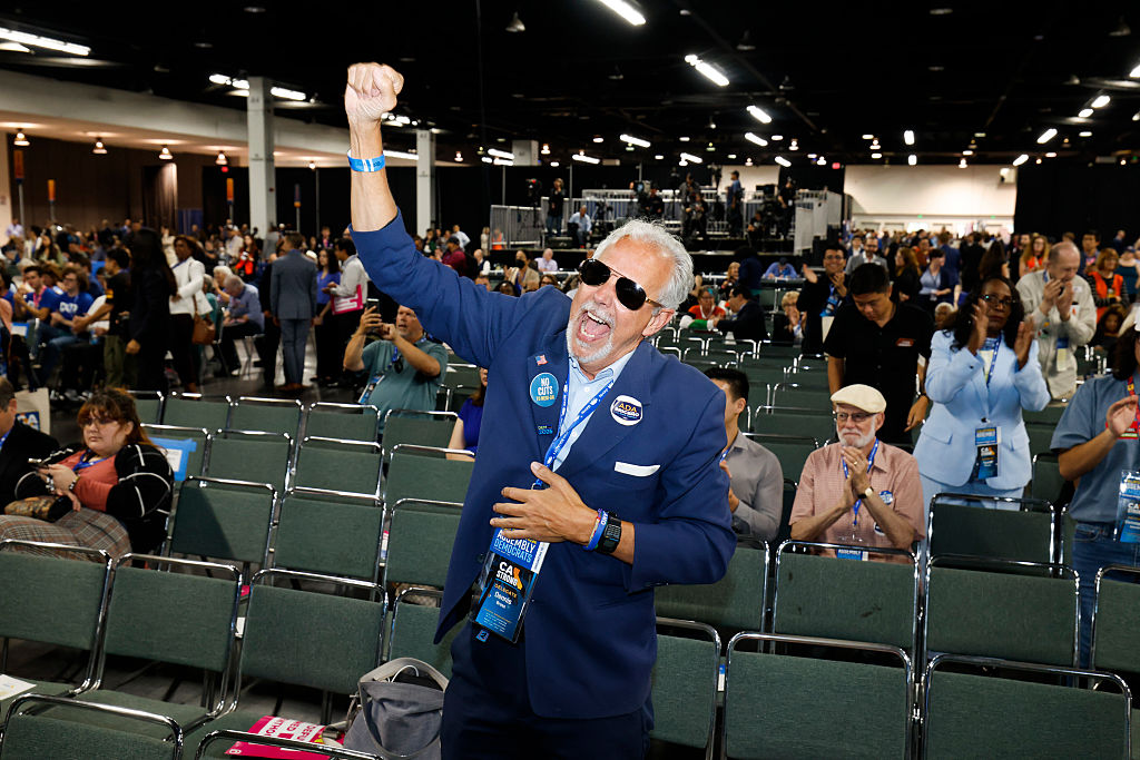 A man in a blue suit and sunglasses enthusiastically raises his arm, surrounded by a seated crowd in a large indoor event space.