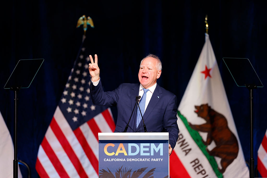 A man in a suit speaks at a podium labeled &quot;CADEM California Democratic Party,&quot; gesturing with two fingers raised. Flags of the USA and California are in the background.