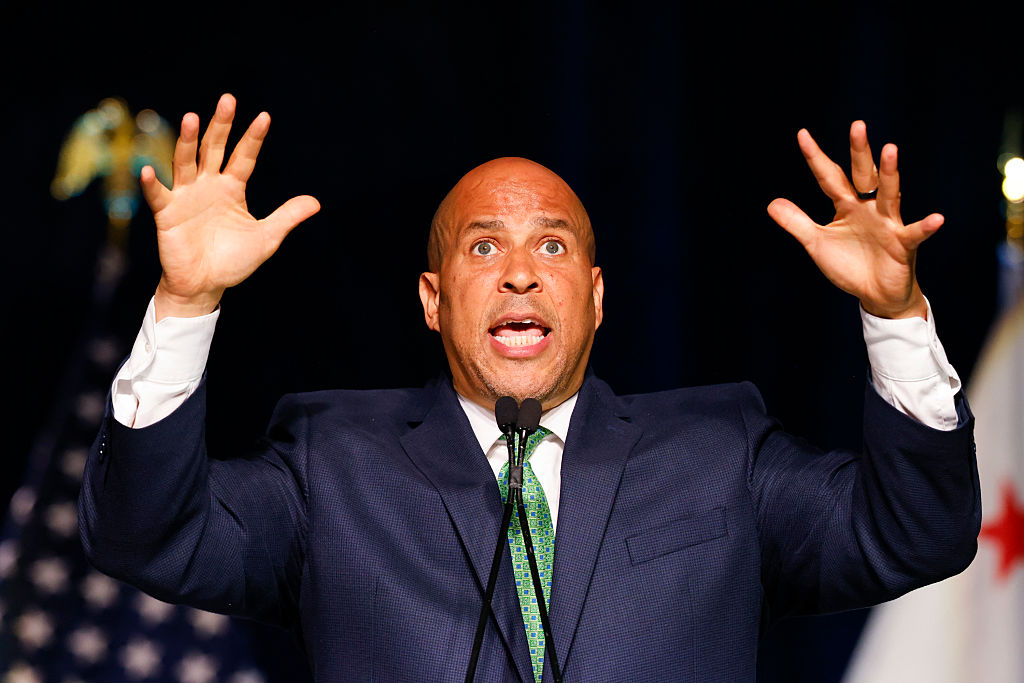 A man in a suit is speaking passionately at a podium, hands raised. The background features blurred flags, including part of the U.S. flag.
