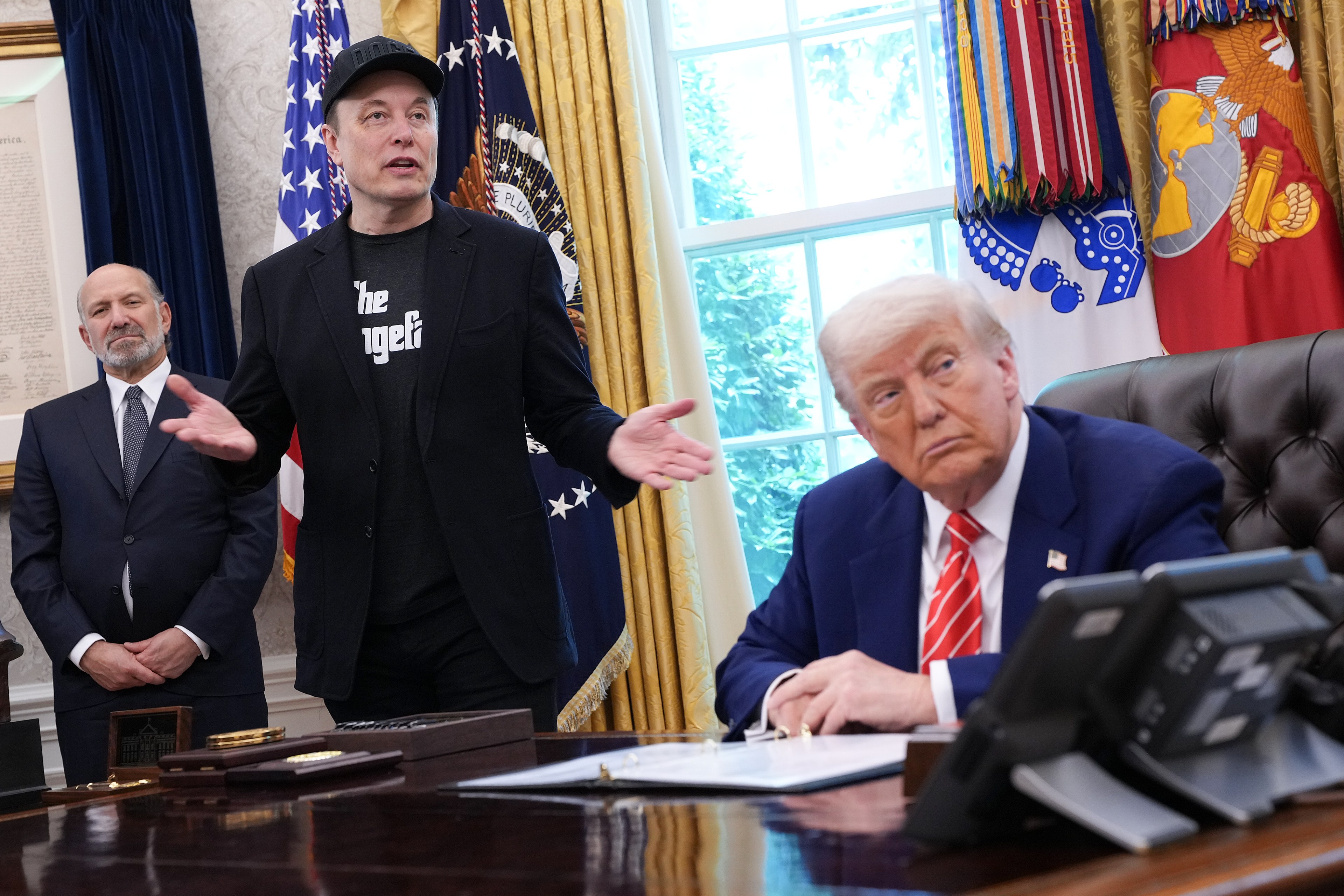 A man in a suit is seated at a desk, listening attentively to another man wearing a cap and casual attire, who stands gesturing with his hands.