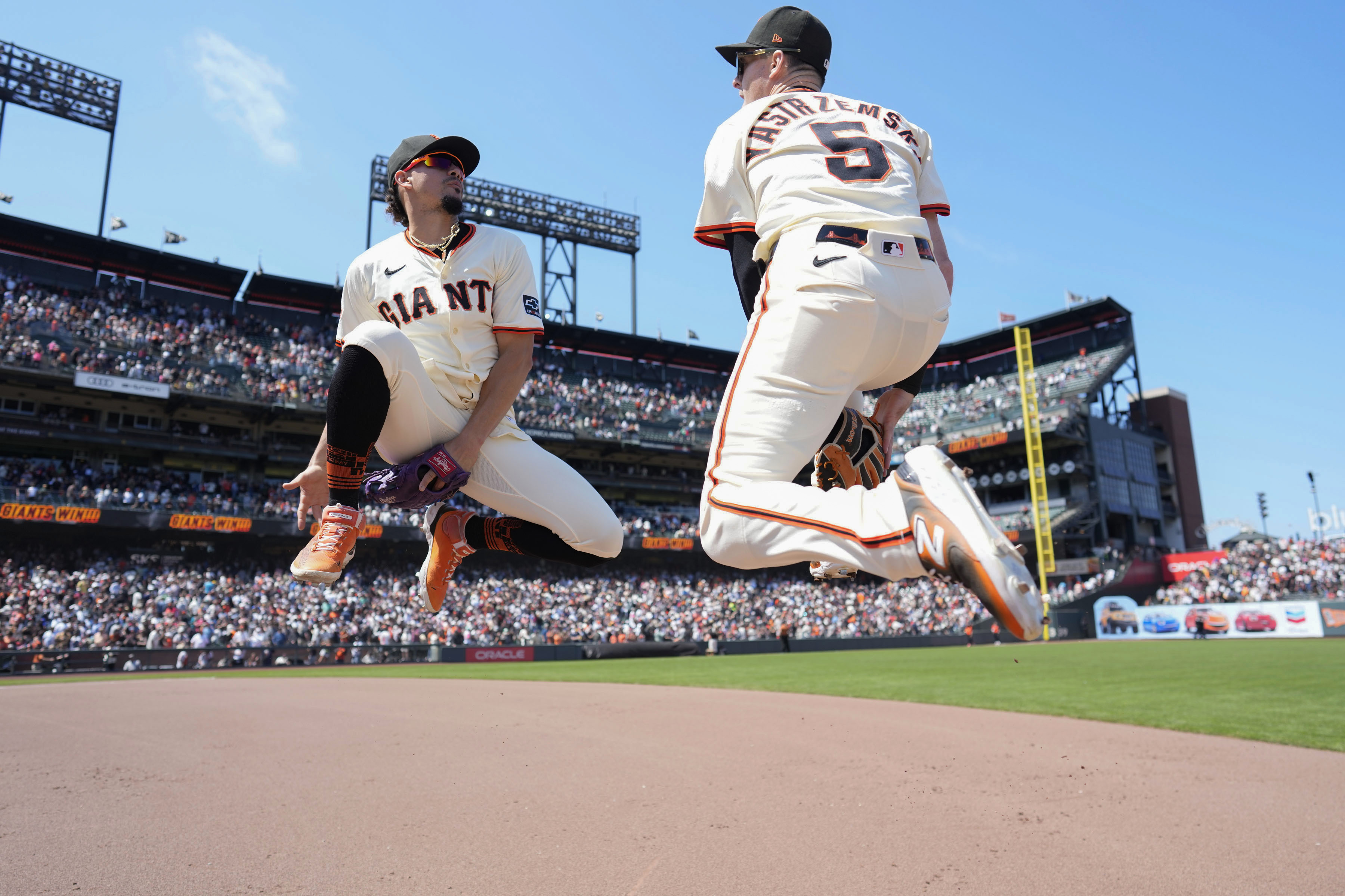 Two baseball players in white uniforms jump and celebrate on a field. The crowd fills the stands under a clear blue sky, with a scoreboard visible.