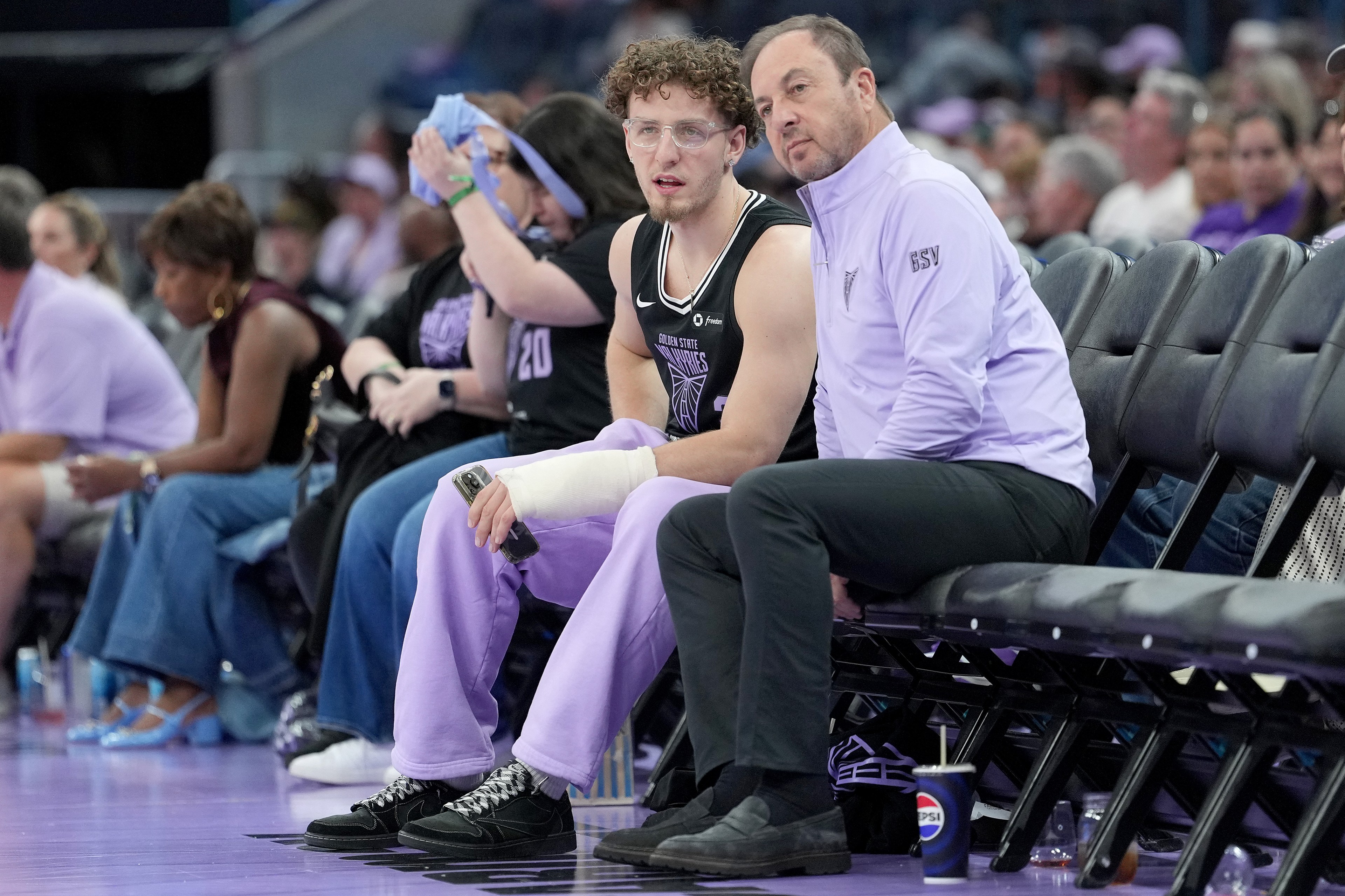 A basketball player with a cast on his arm sits beside a coach on the bench. They're surrounded by spectators, some wearing purple. A Pepsi cup is on the floor.
