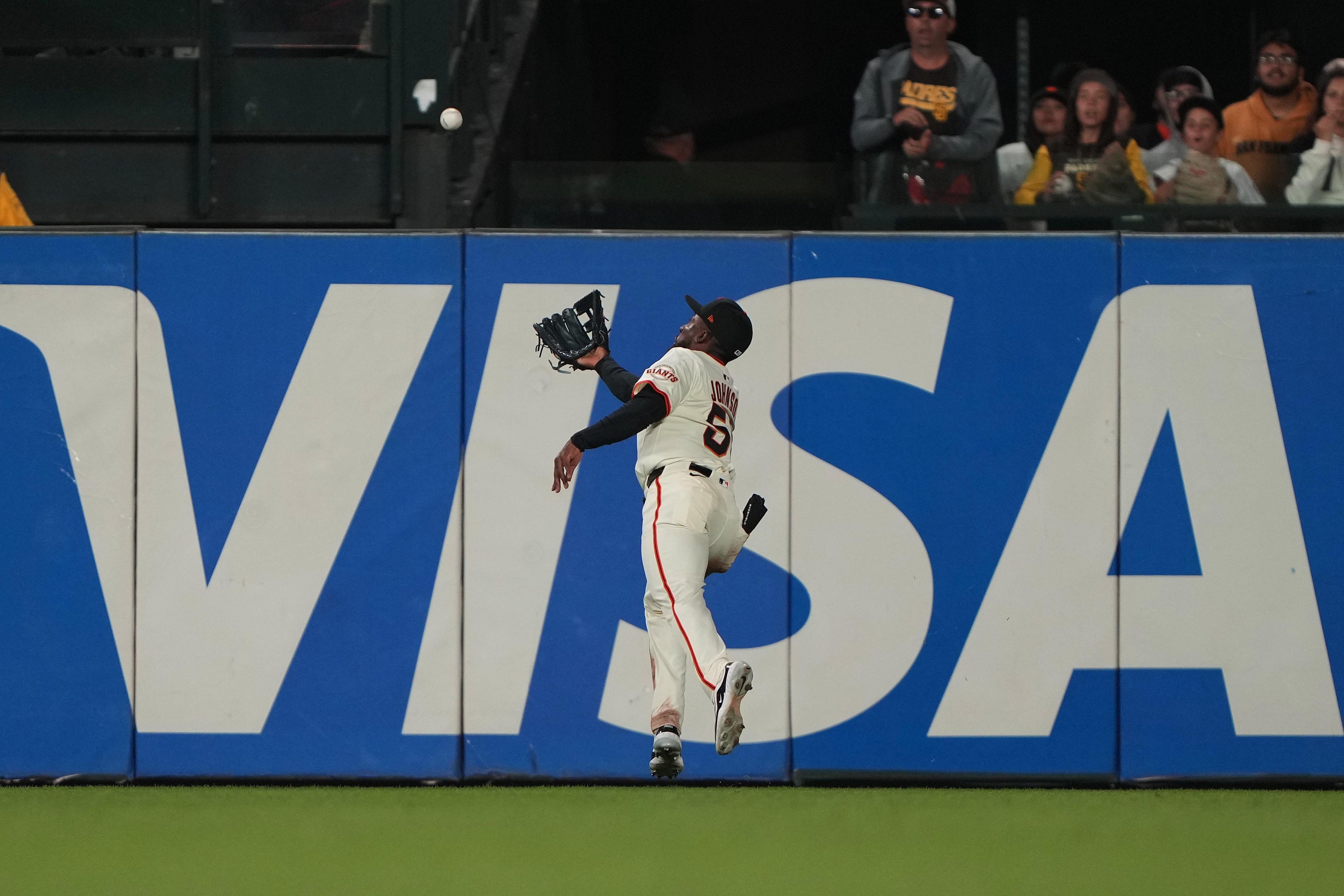 A baseball player in a white uniform leaps in the air near a wall with a &quot;VISA&quot; ad, attempting to catch a ball. Spectators watch from the stands behind.
