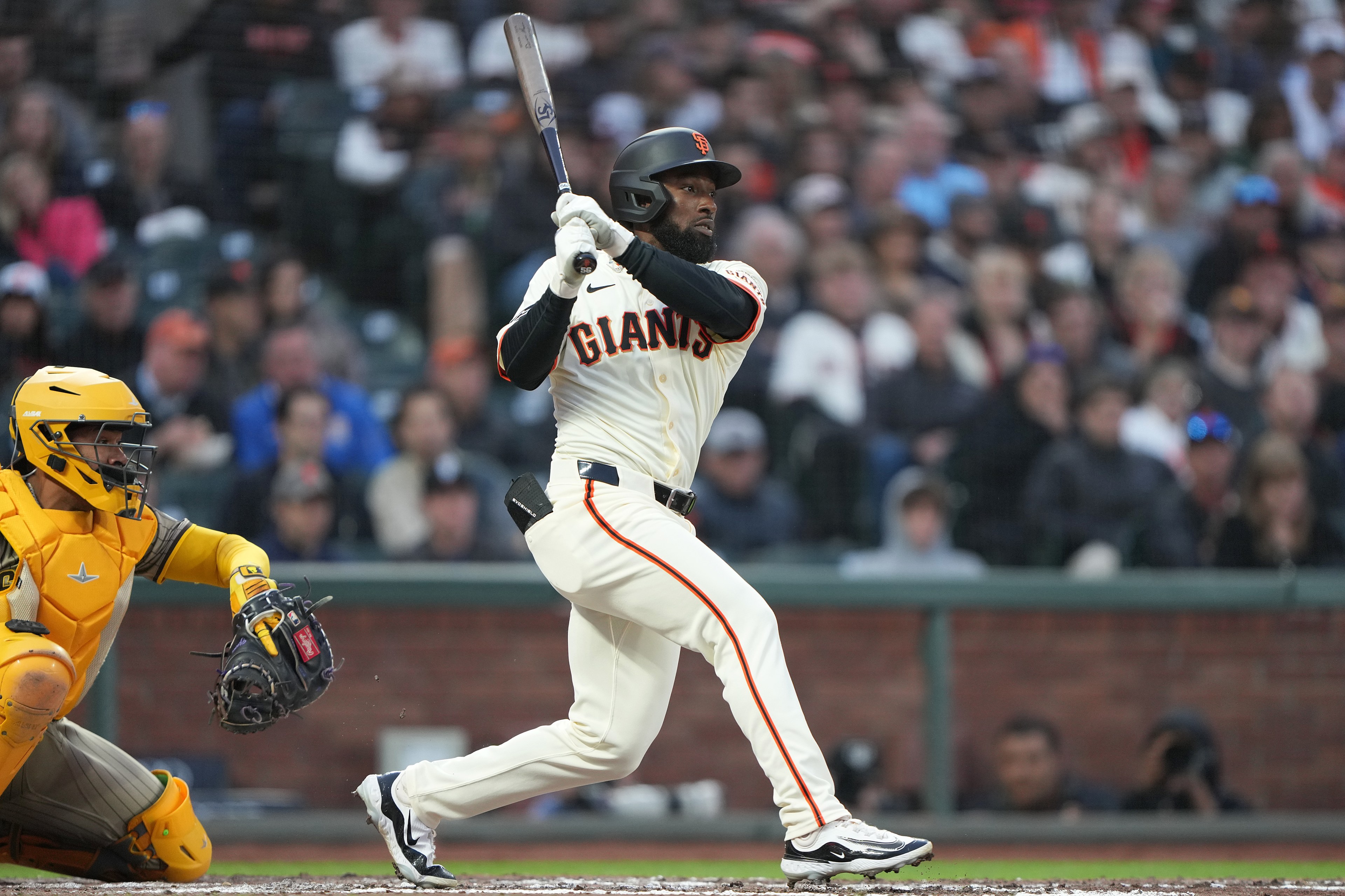 A baseball player in a white Giants uniform swings a bat during a game. A catcher in yellow gear is poised to catch the ball. Spectators fill the stands.