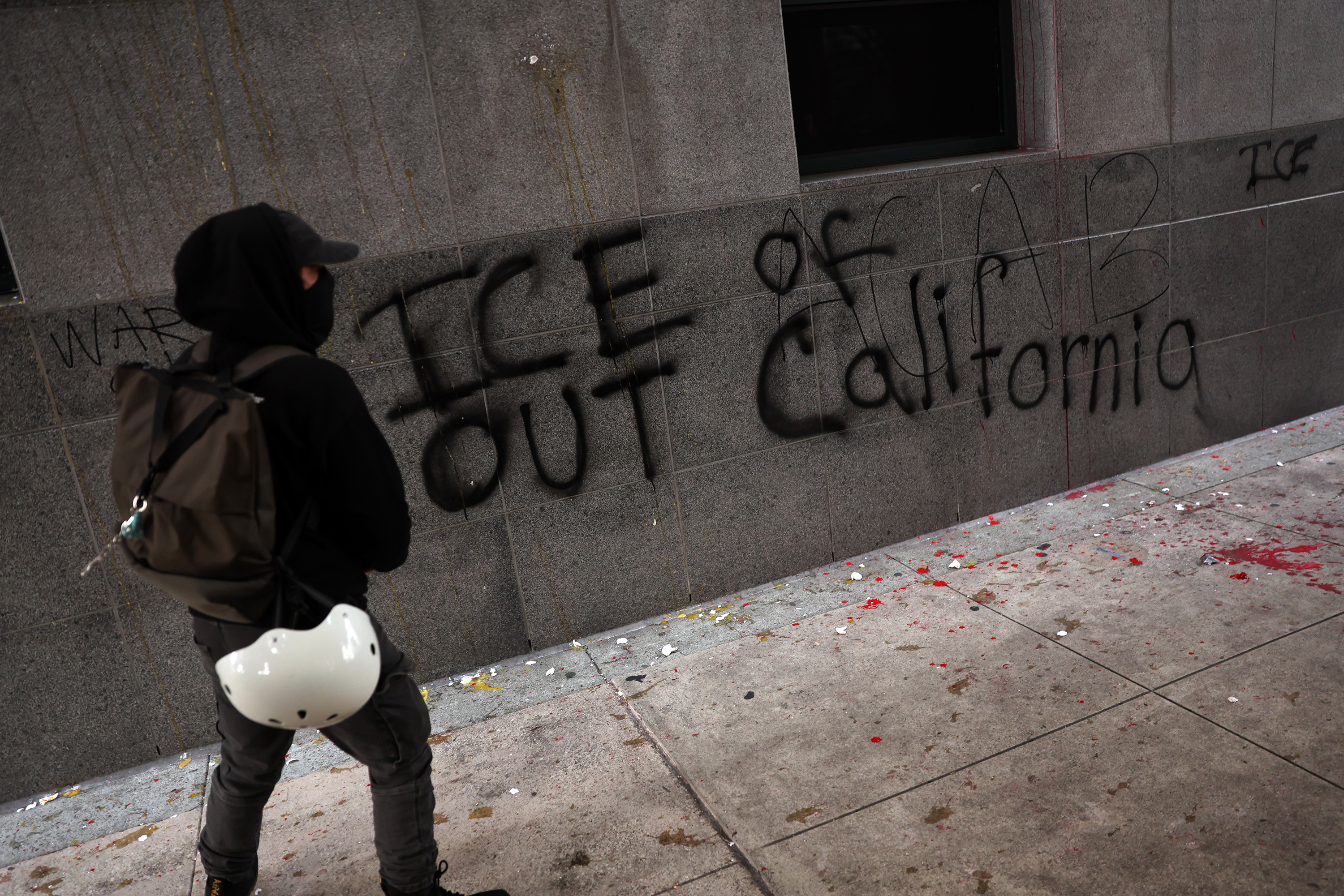 A person in dark clothing and a backpack stands by a graffiti-covered wall reading "ICE OUT of California." The ground has splatters of red and white.