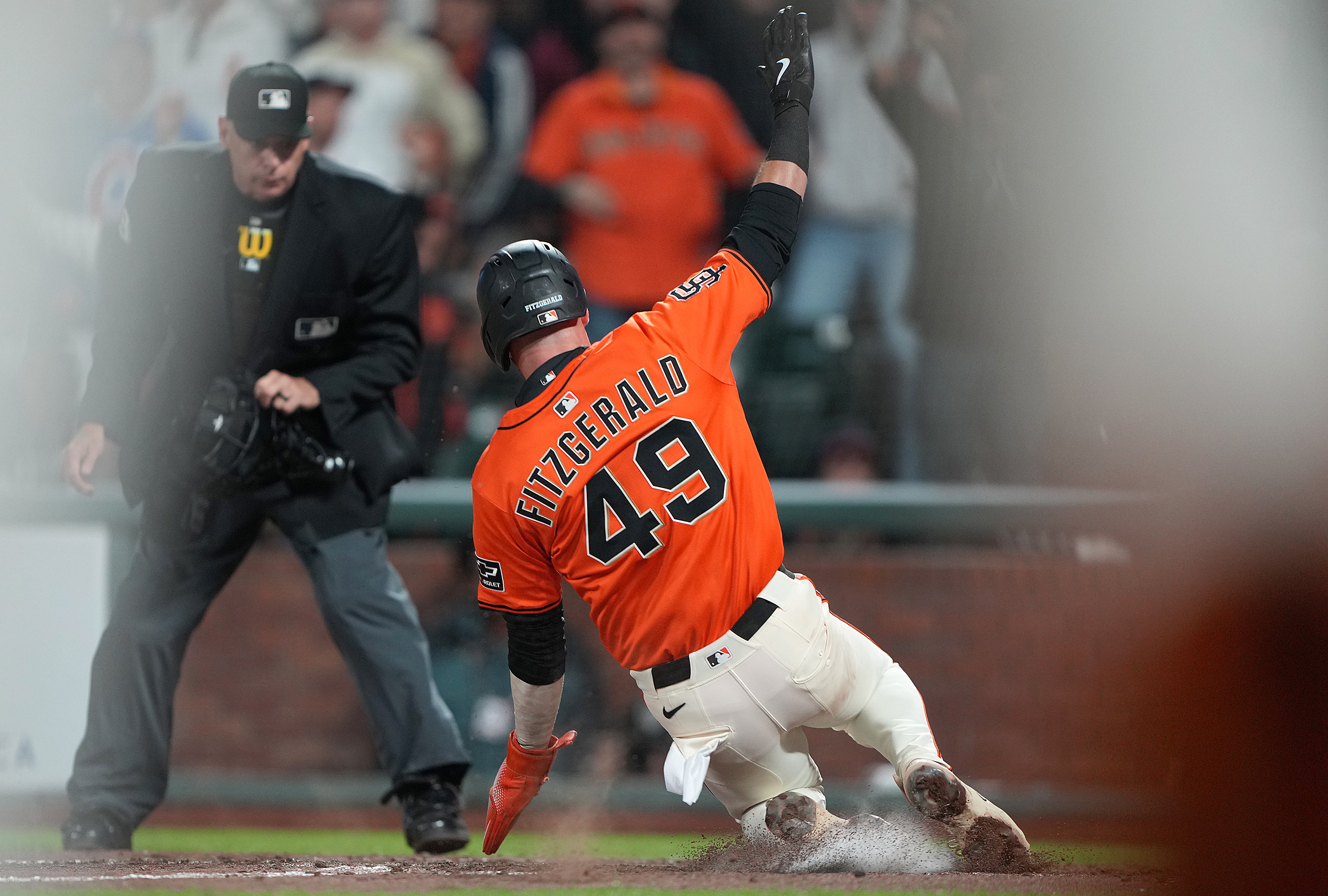 A baseball player in an orange jersey with the number 49 slides into a base. An umpire in black watches closely, and the scene is blurred around the edges.