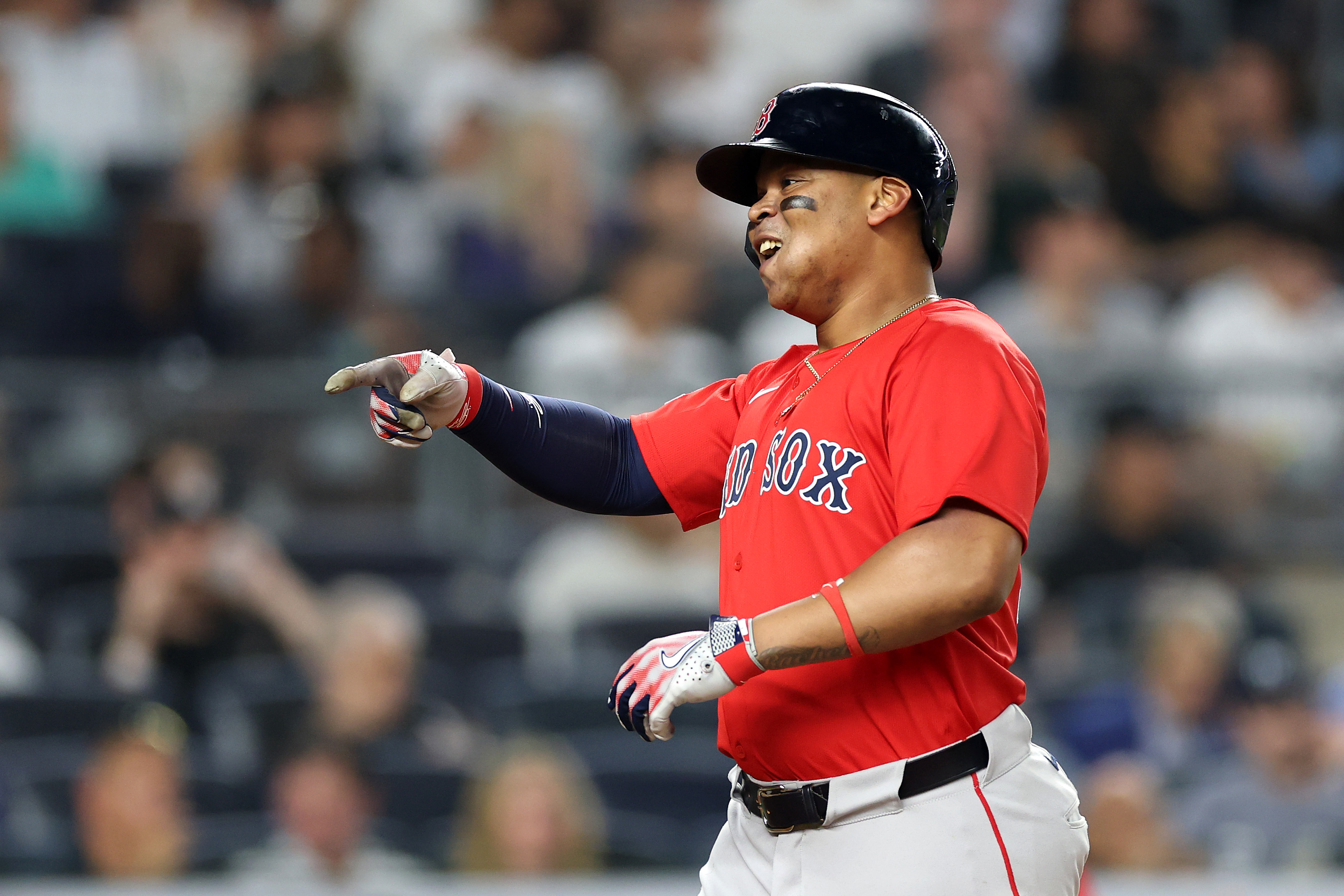 A baseball player in a red jersey and helmet points and smiles on the field, his face marked with black streaks. The blurred crowd is visible in the background.