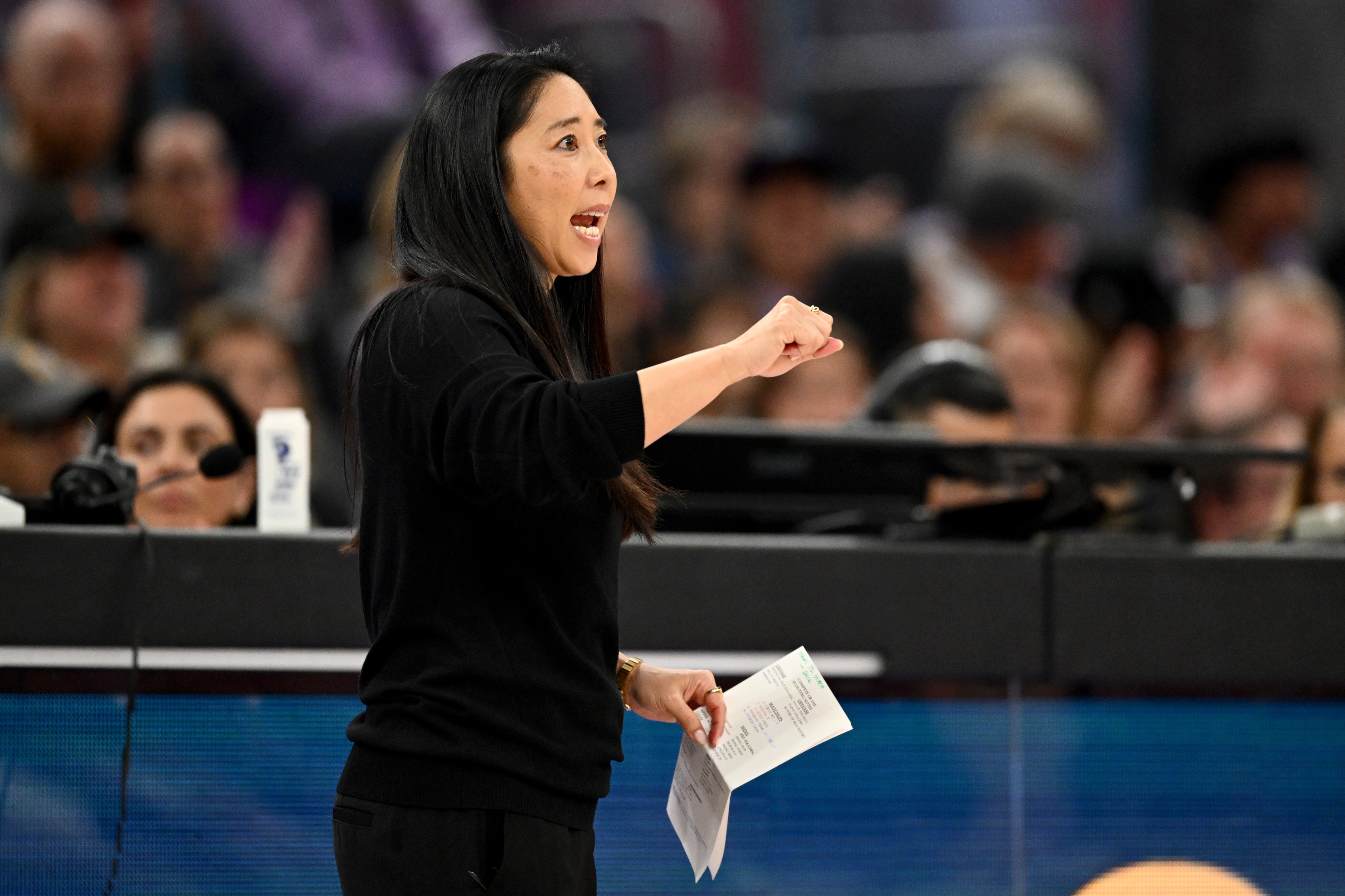 A woman with long dark hair, in a black sweater, is speaking energetically while holding papers. A crowd is in the blurred background.