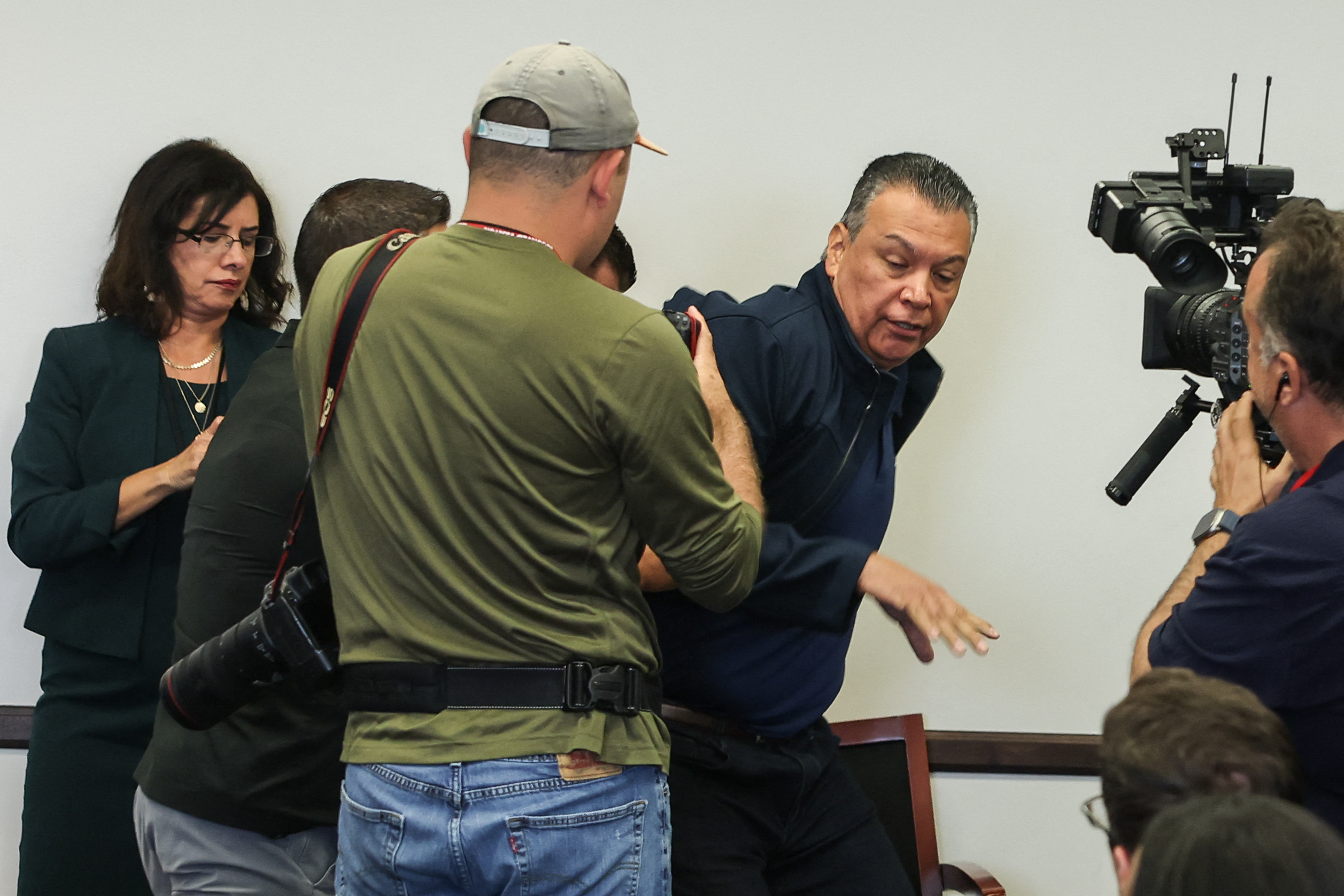 A media scrum captures a man in a navy sweater as he moves past a group, including a photographer in a cap with a camera and another person in a green jacket.