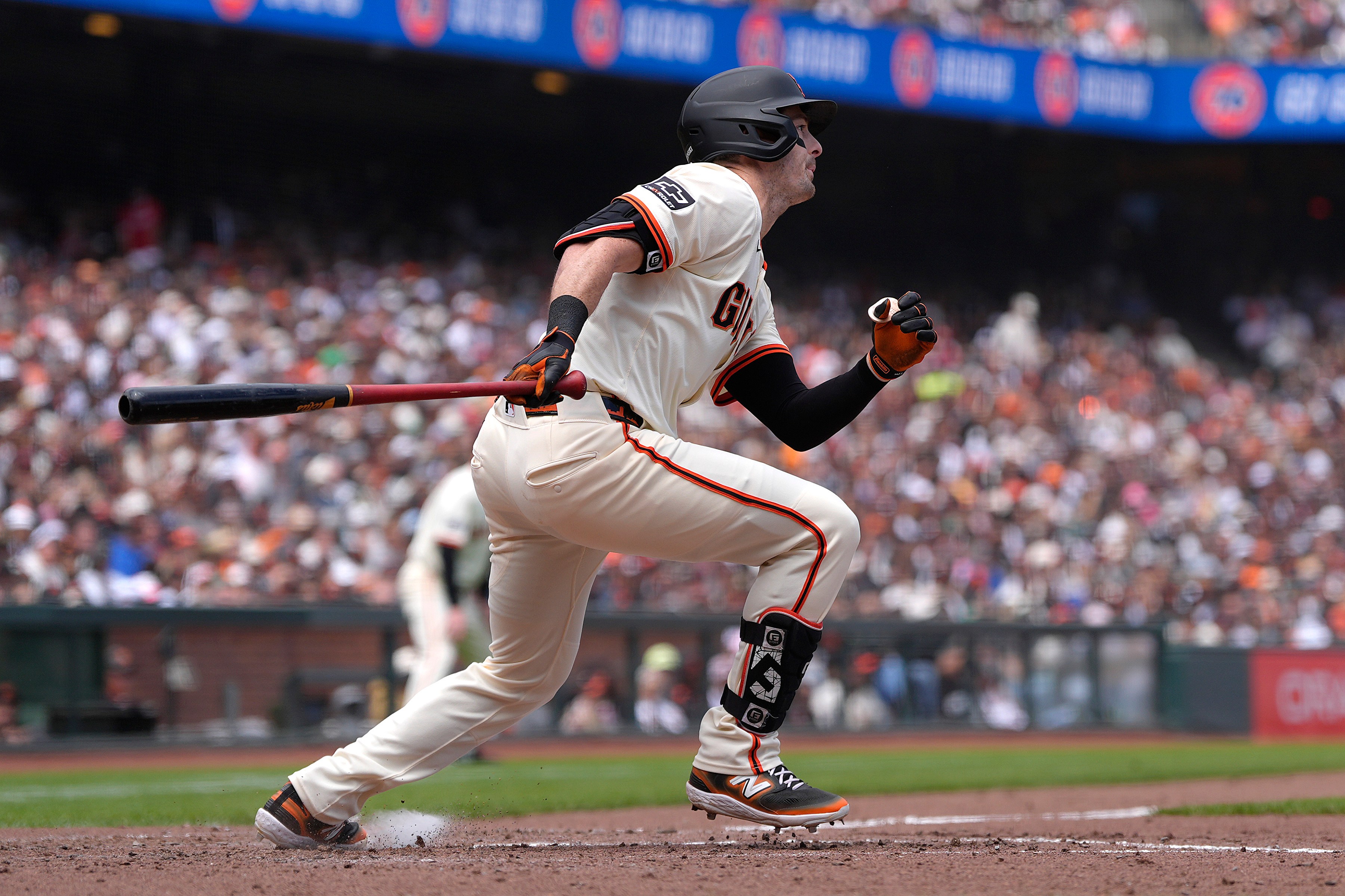 A baseball player in a white and orange uniform is running, gripping a bat. The crowd in the background is blurred, suggesting a lively ballpark scene.