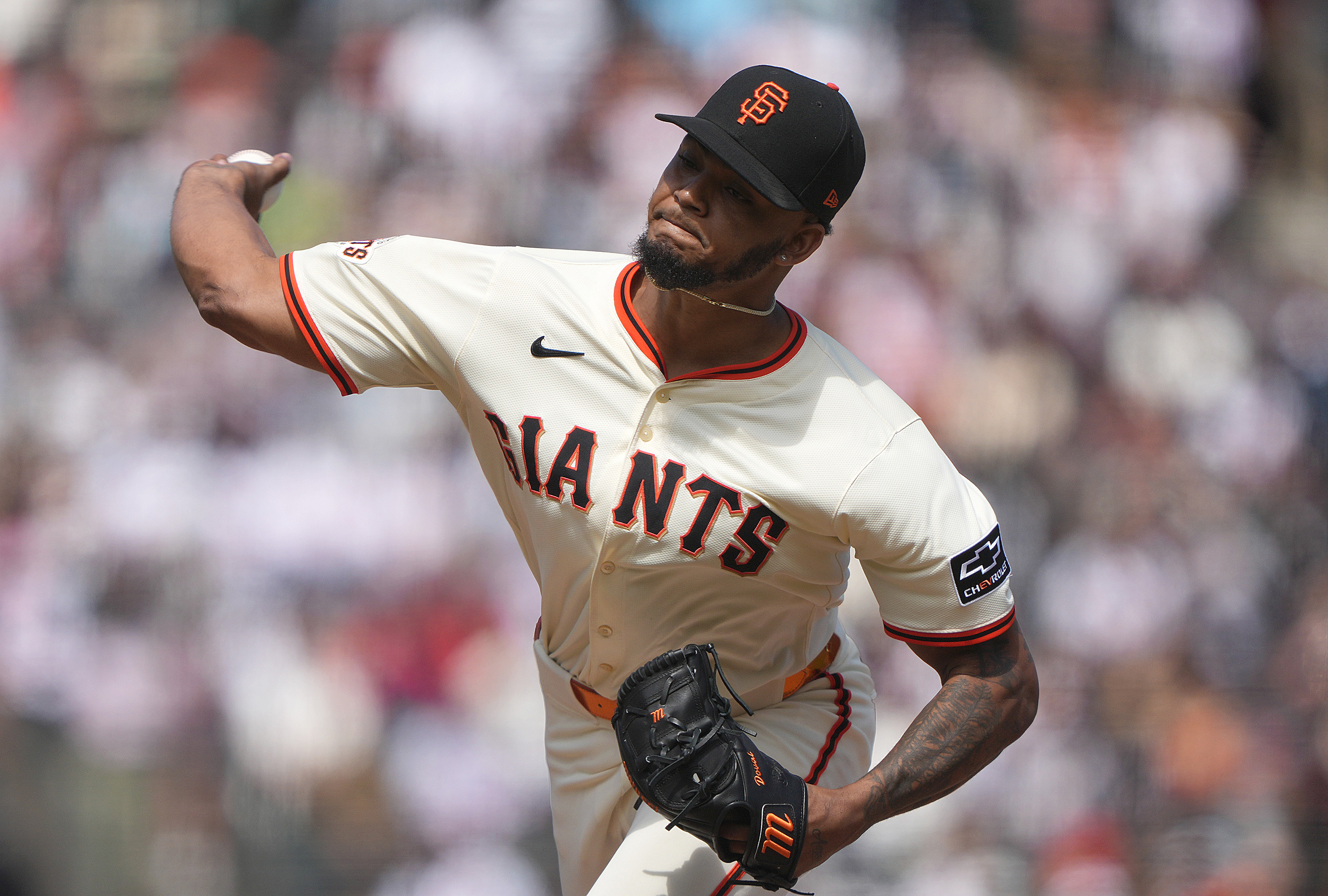 A baseball player wearing a Giants uniform is in a pitching motion. He’s in a stadium, holding a ball, with a focused expression and a black cap.