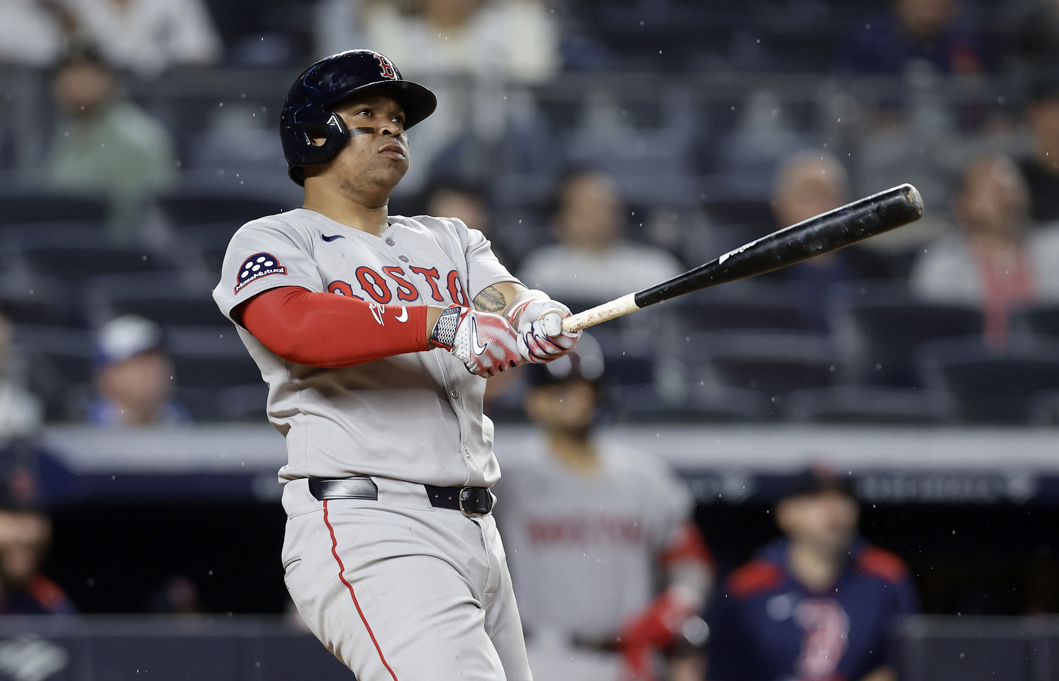 A baseball player in a gray &quot;Boston&quot; uniform follows through on a swing, eyes focused on the ball. The stadium seats in the background are blurred.