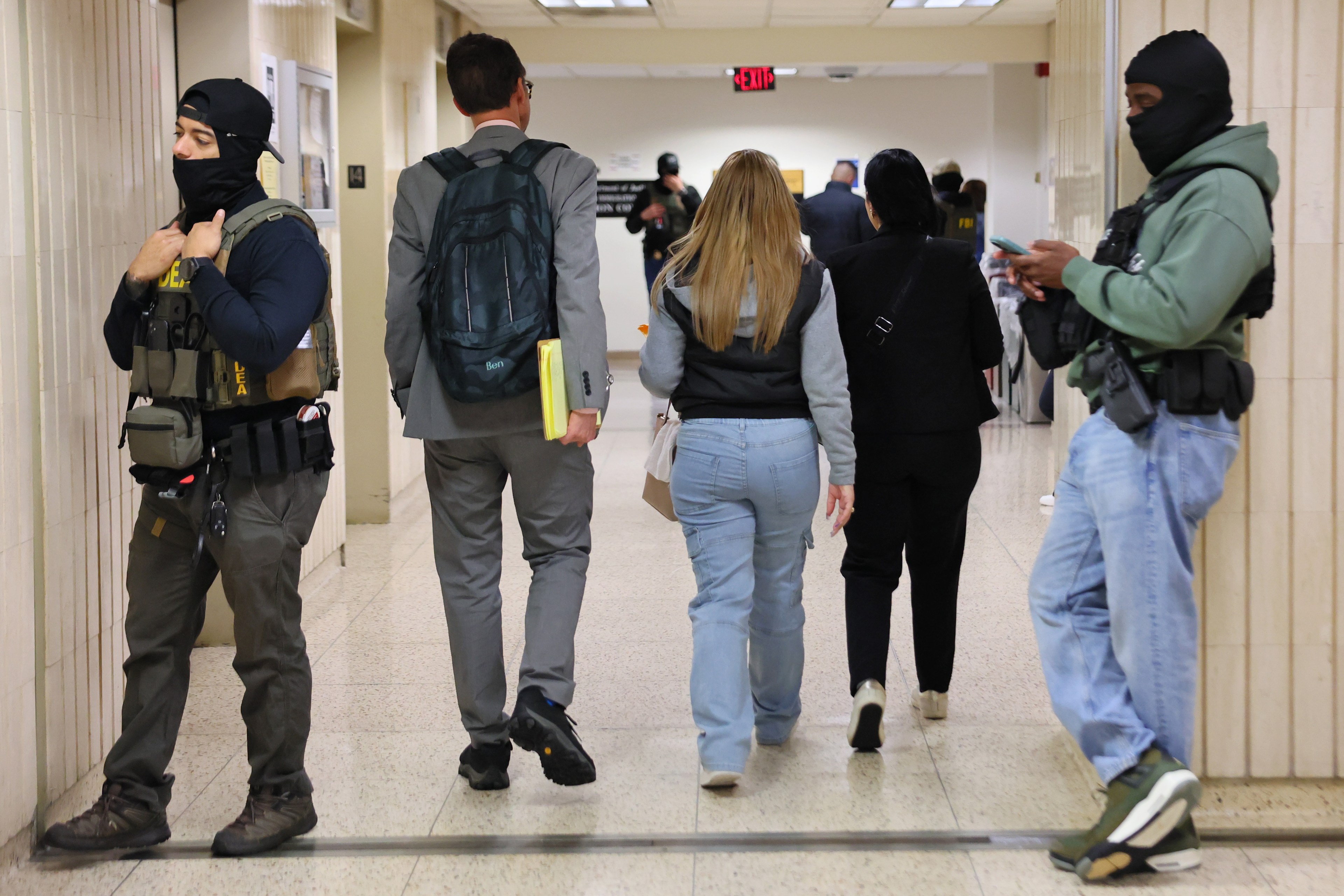 Two masked officers stand guard in a hallway as several people walk away, including a person in a suit and another wearing casual clothing, carrying items.