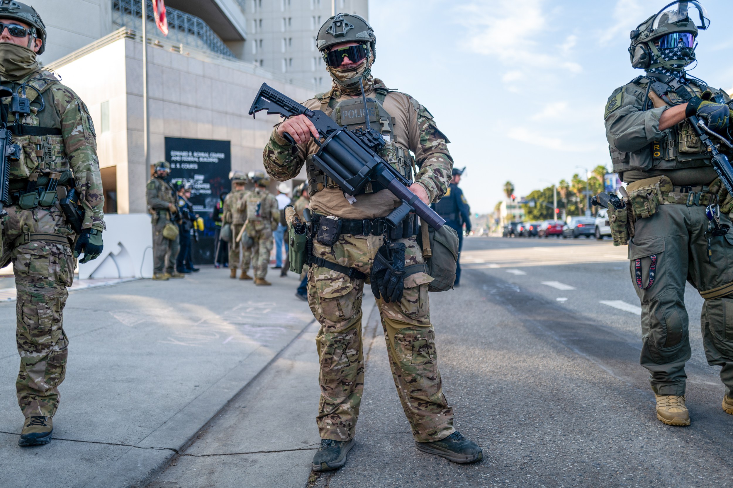 Three armed individuals in tactical gear stand on a street, holding large weapons. They appear to be part of a security detail or law enforcement.