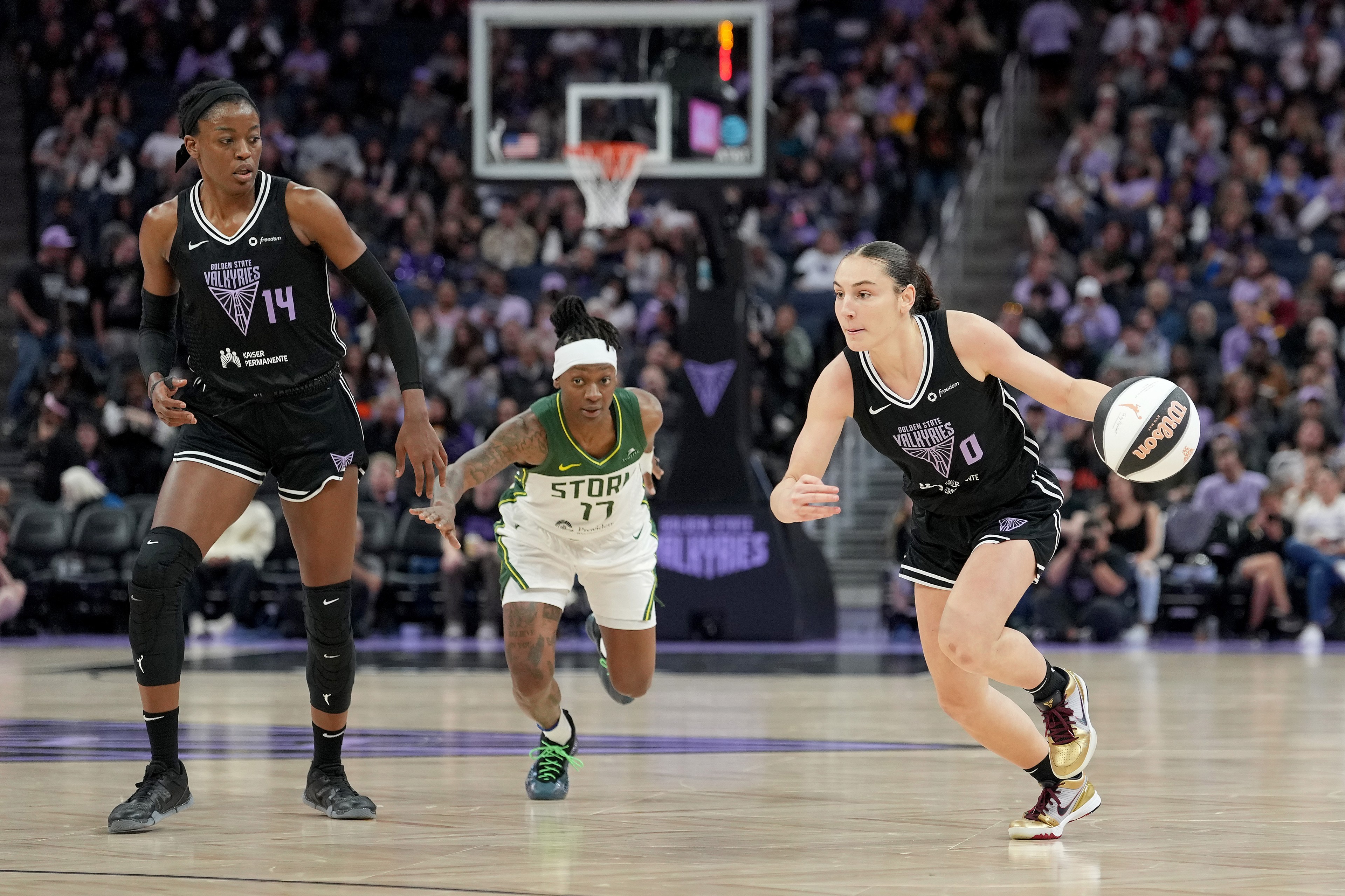 A basketball player in black dribbles the ball while a teammate and an opponent in white chase her on the court, with the crowd visible in the background.