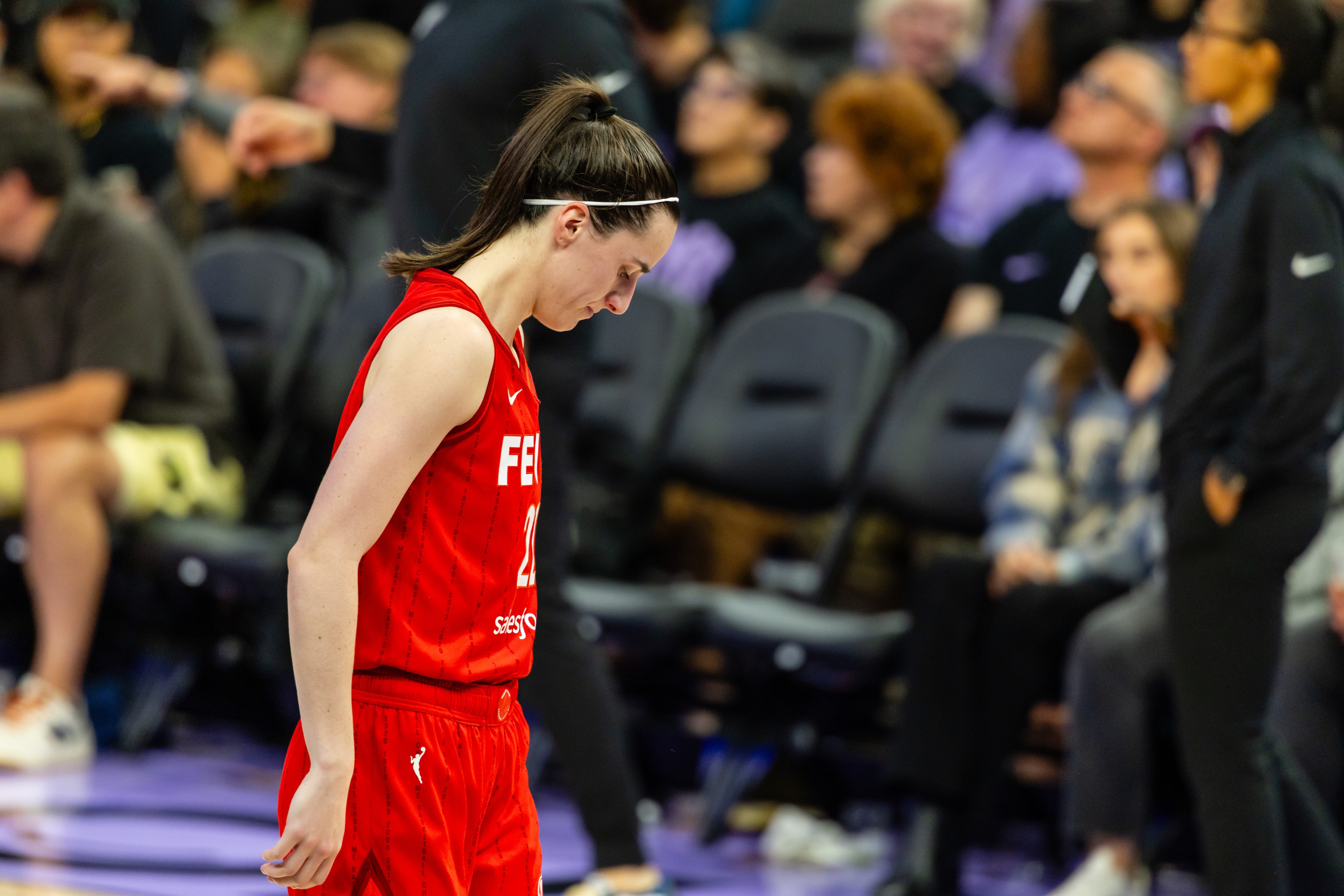 A basketball player in a red uniform walks with a focused expression. The background shows a blurred audience watching the game.